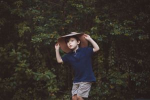 Young boy in navy polo shirt holding wide brim hat standing in front of lush green foliage.