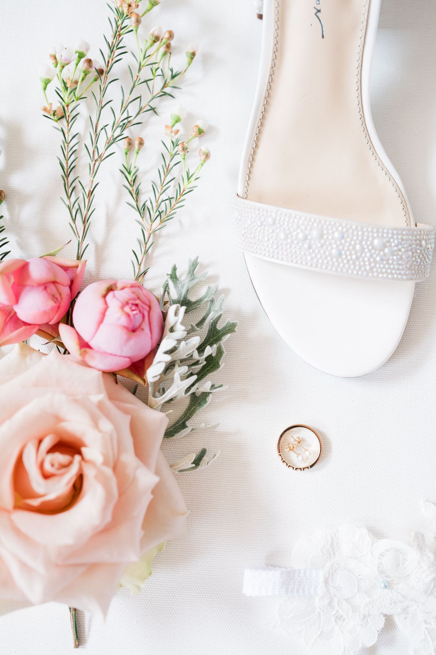 Wedding details flatlay with pink roses, delicate white shoes, and a wedding ring on white background.