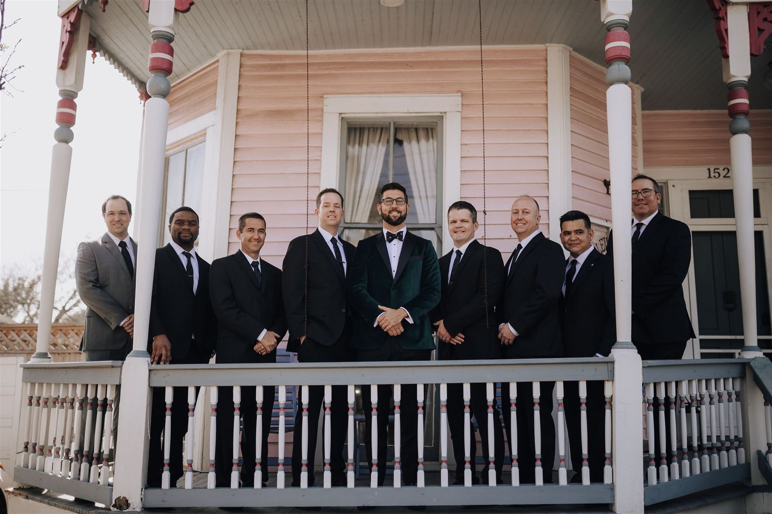 Groomsmen in dark suits pose together on a vintage pink house porch.