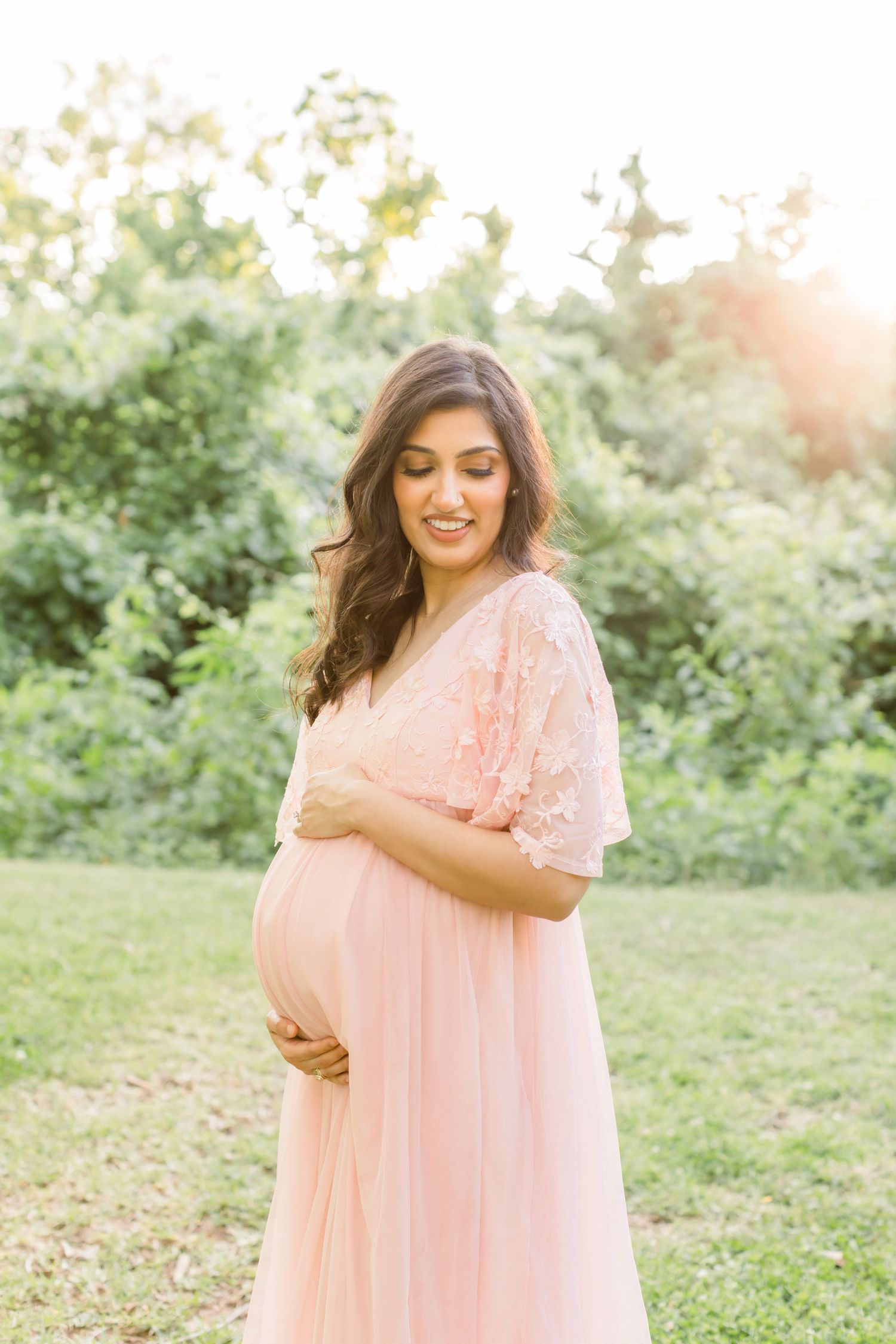 A radiant maternity portrait of a woman in a blush pink dress amid greenery.