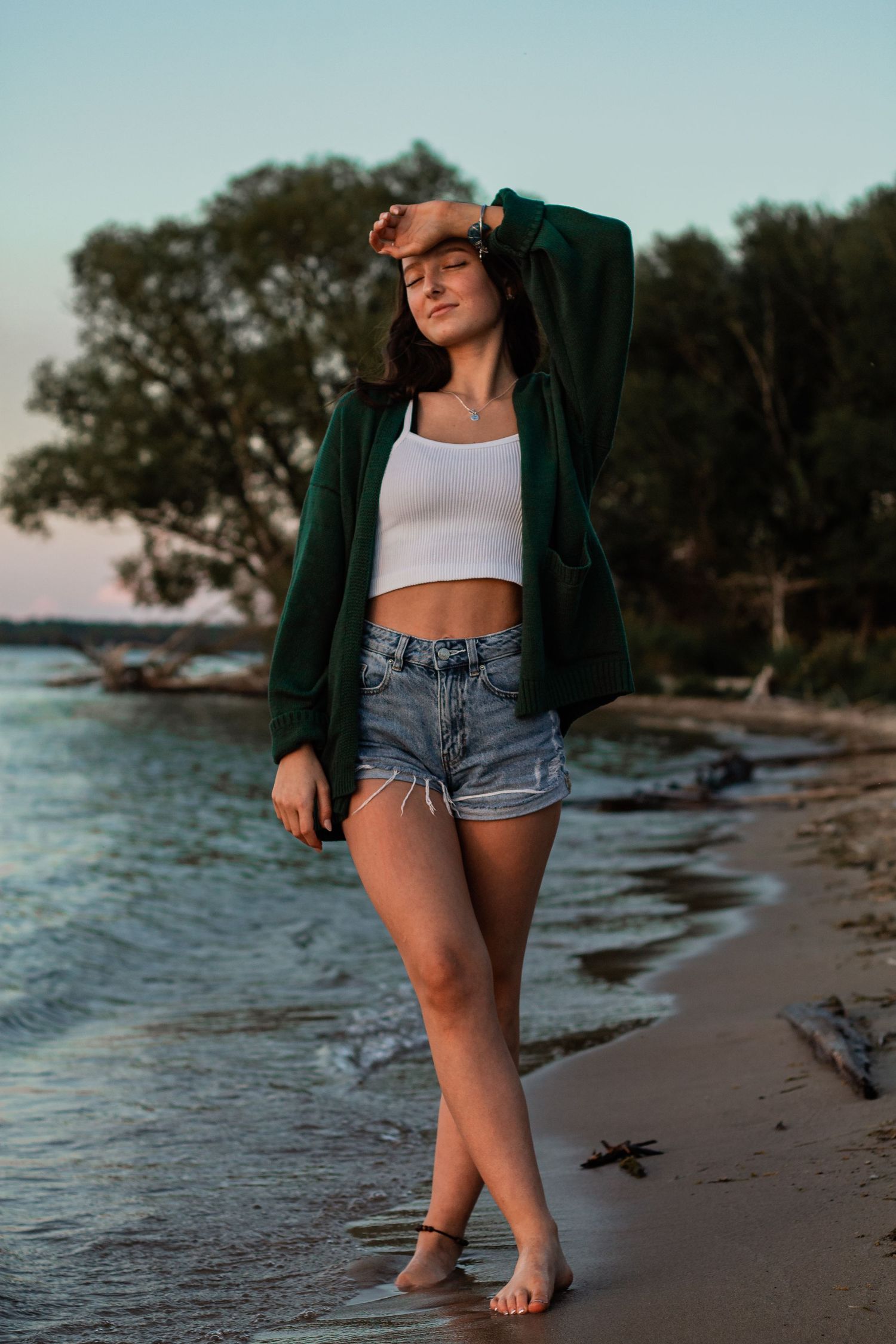Casual beach style with green cardigan and denim shorts along waterfront at dusk.