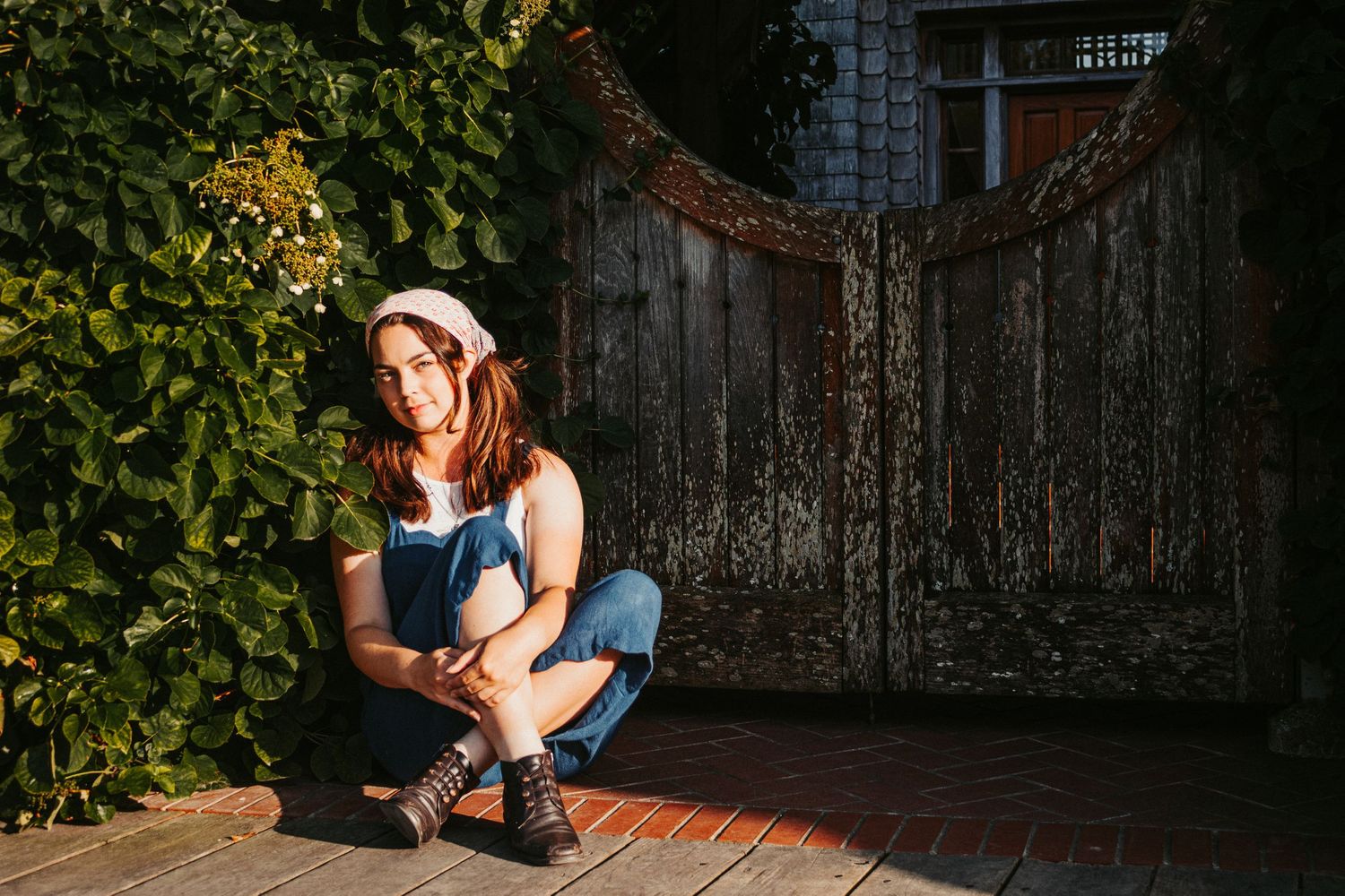 Casual pose by a vintage wooden gate surrounded by green leaves and brick pathway.