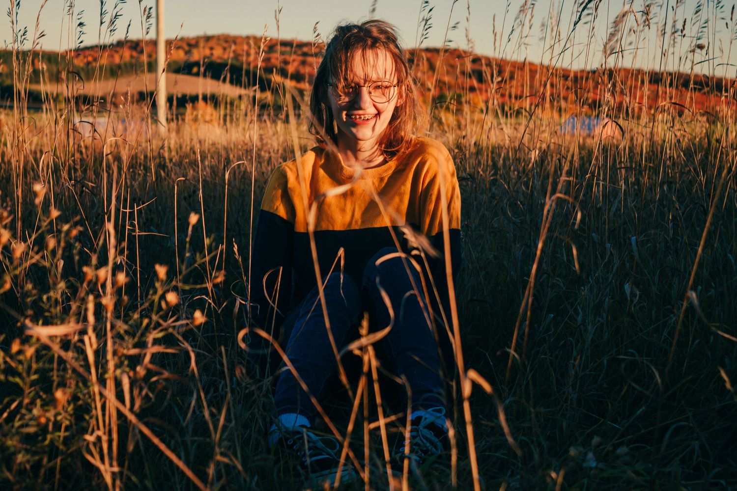Outdoor portrait in orange hoodie among tall grass during sunset with hills in background.