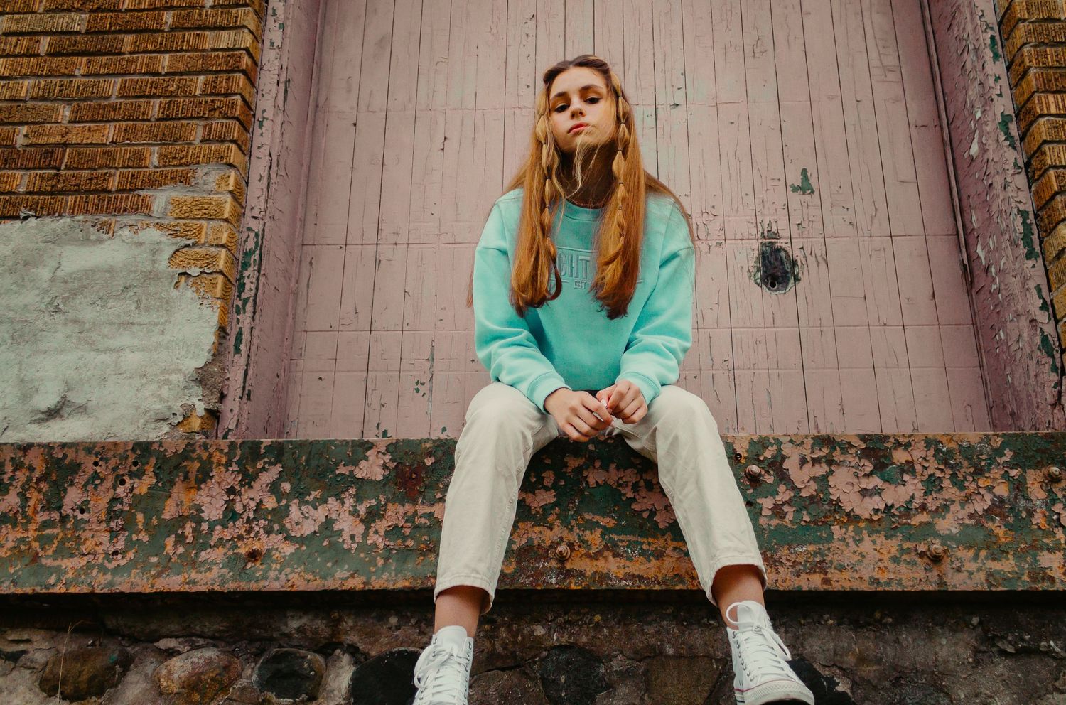 Lifestyle portrait of woman in turquoise sweatshirt and white pants sitting against pink rustic door.