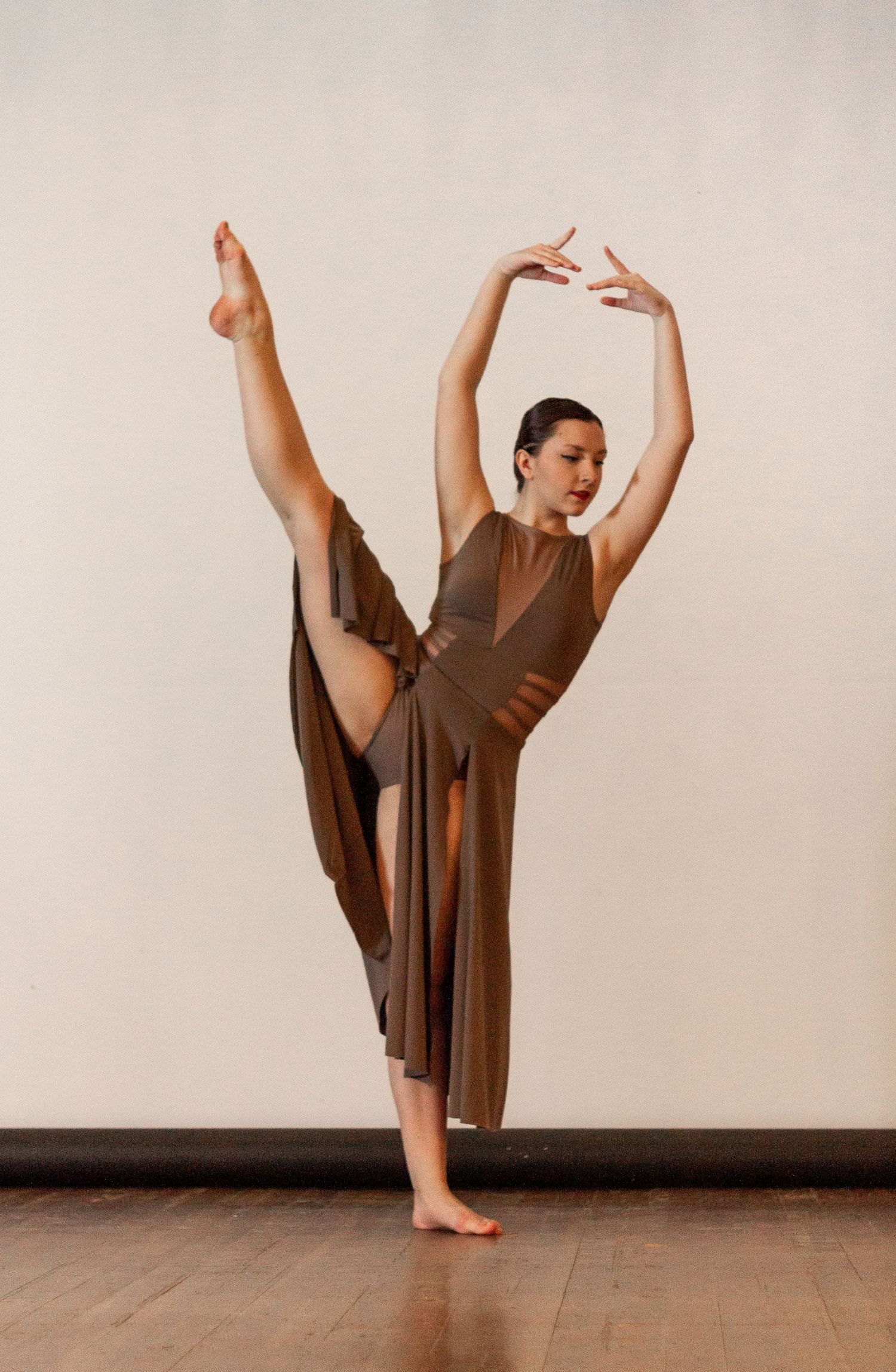 A graceful dancer in a brown dress performs a dramatic split pose against a neutral backdrop.