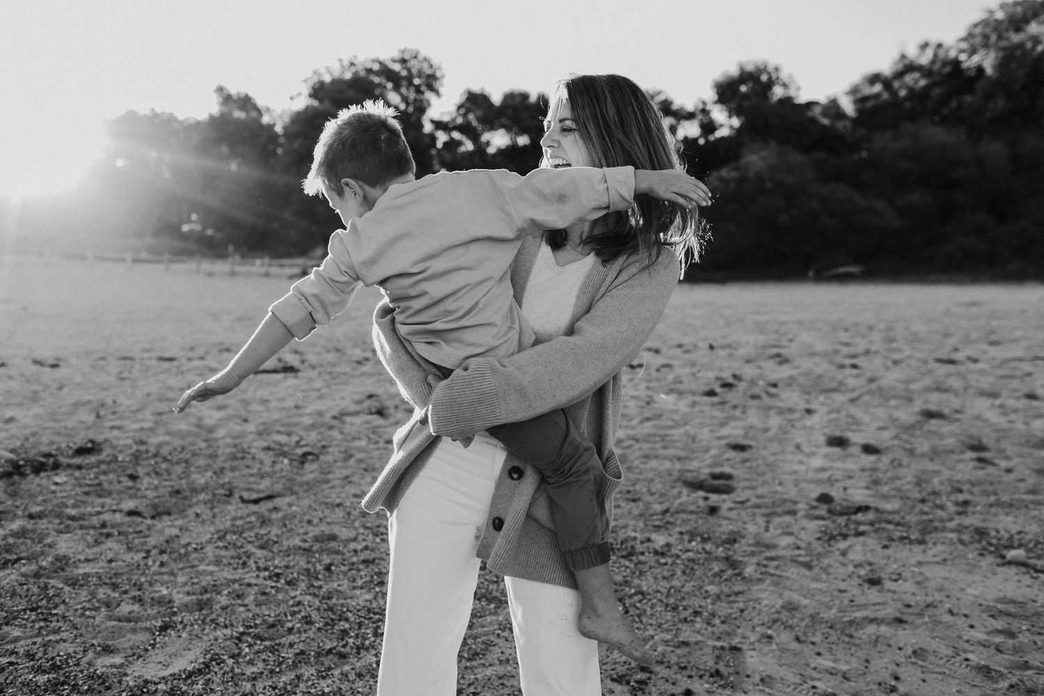 Mother joyfully lifts child on sunny beach in black and white candid family photo.