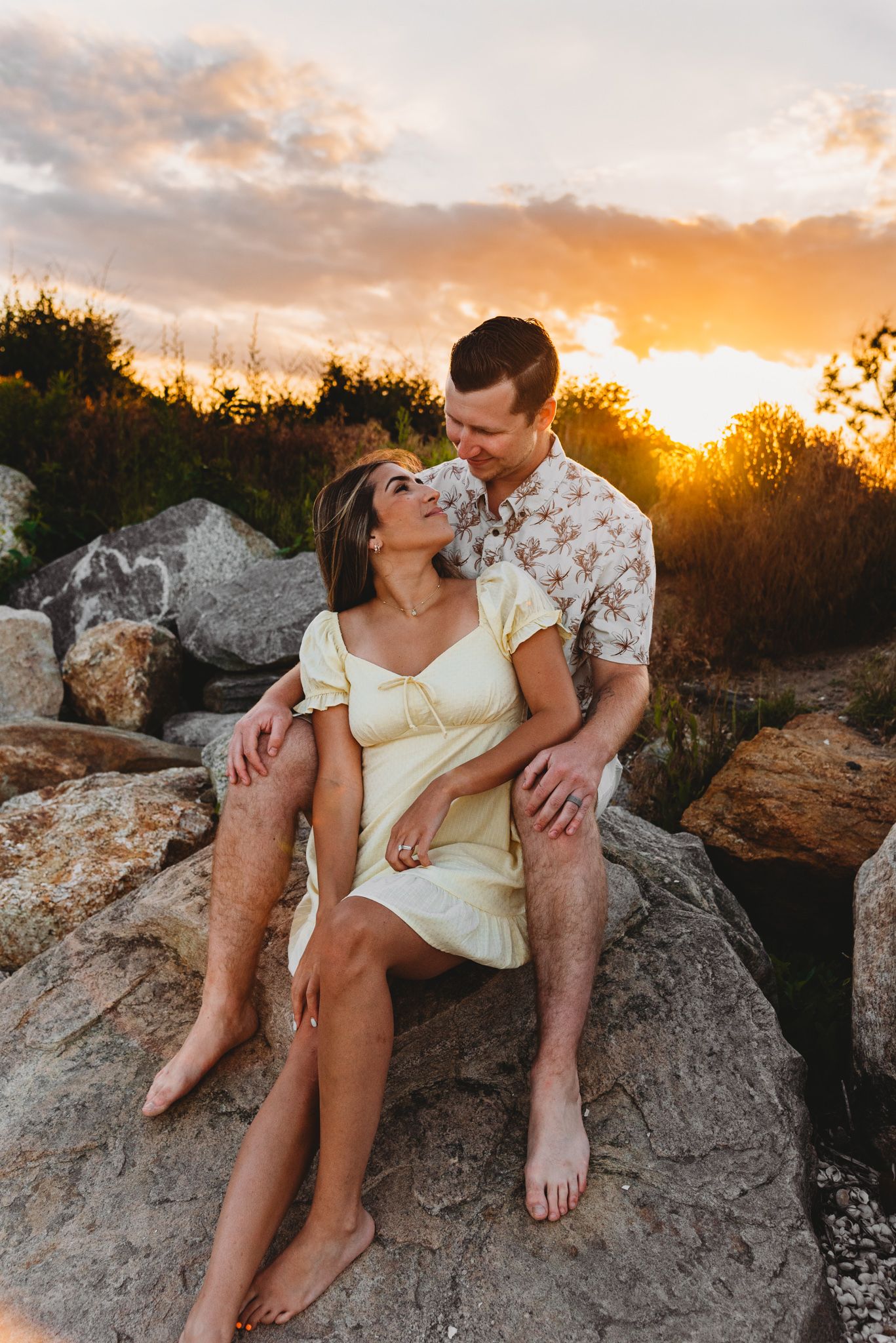 Romantic couple embraces on rocky outcrop during golden sunset wearing casual summer attire.