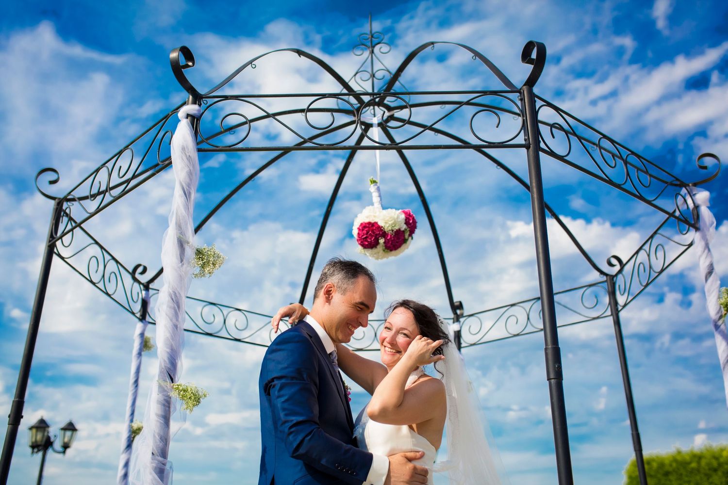 Couple shares intimate moment under decorative metal gazebo against blue sky.