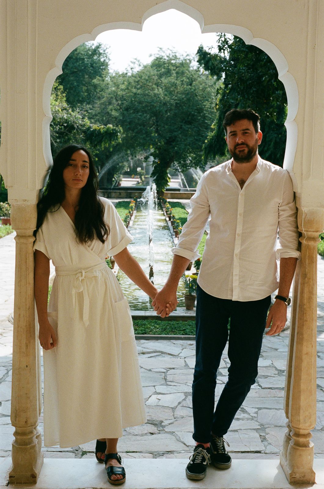 A couple in white outfits holds hands while standing under an ornate archway in a garden setting.