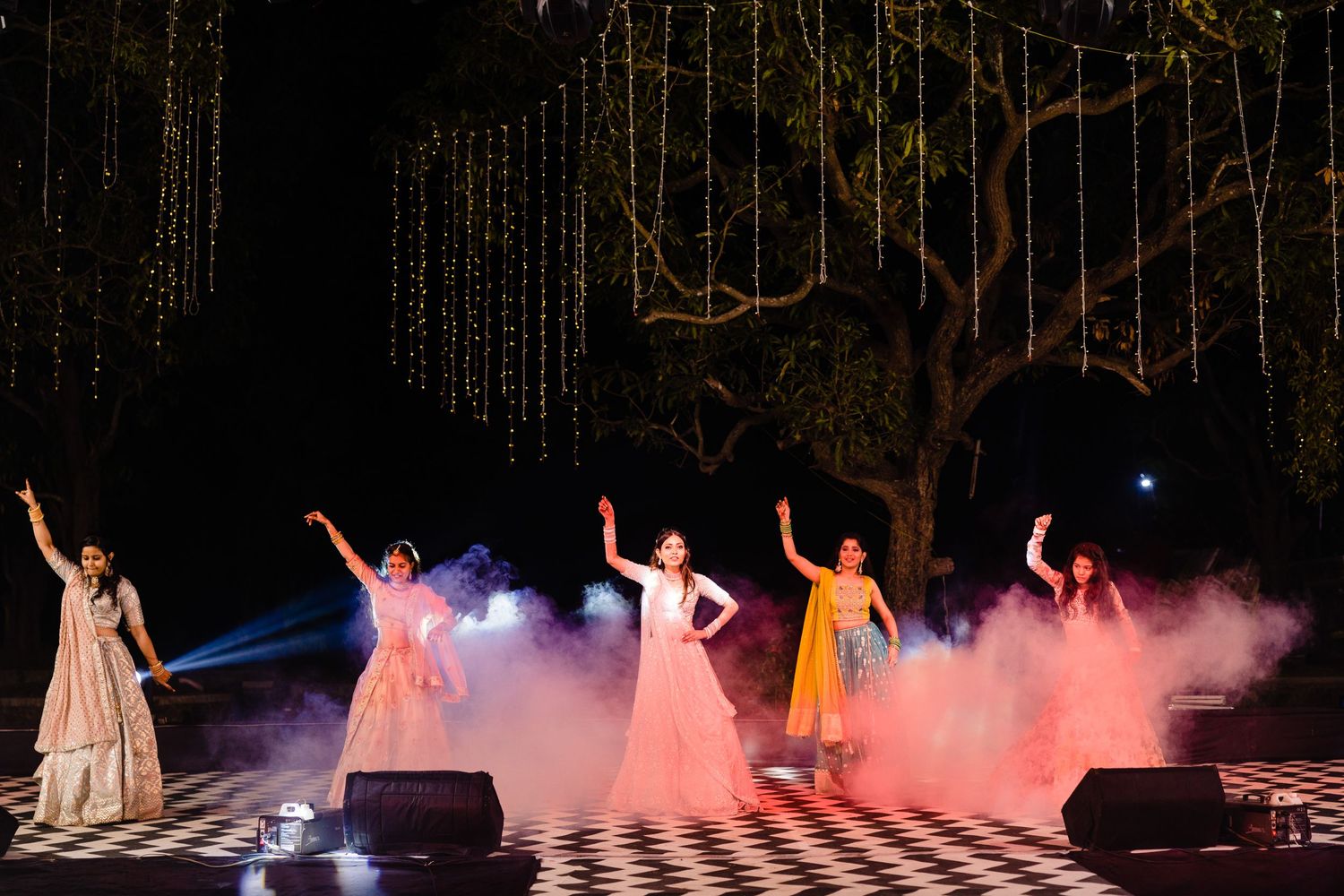 Theater performers in elegant costumes dancing on a checkered stage floor with dramatic pink lighting effects.