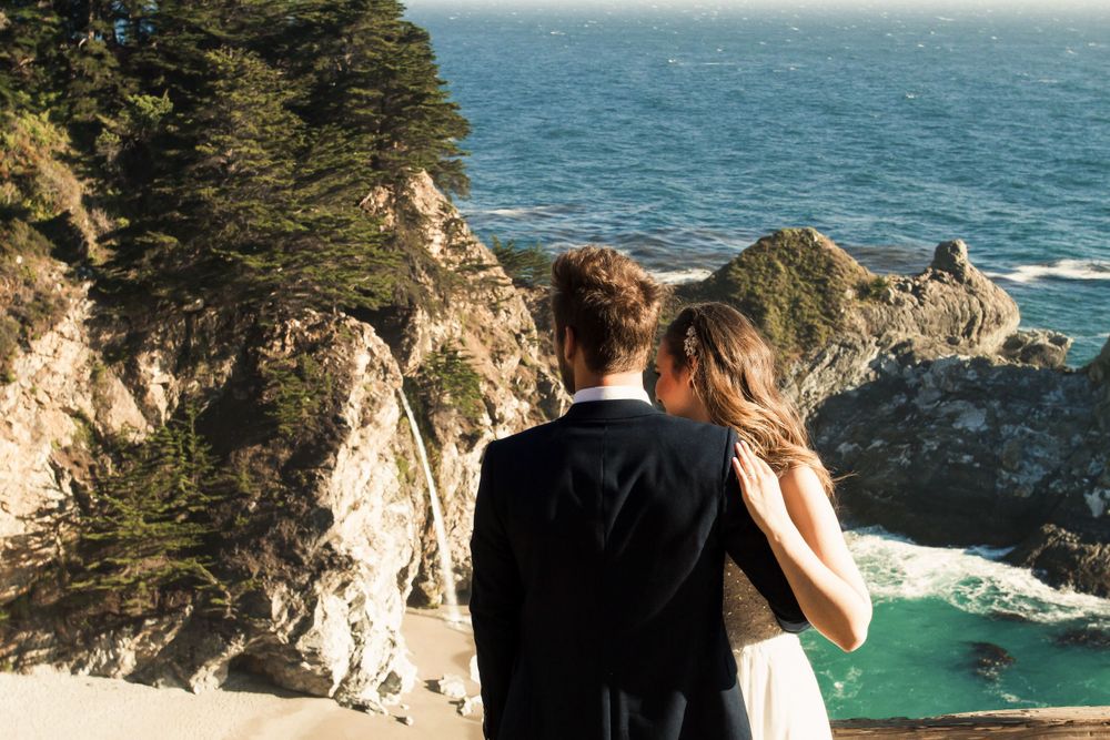 Two people admiring dramatic coastal views of Big Sur from a clifftop overlook above turquoise waters.
