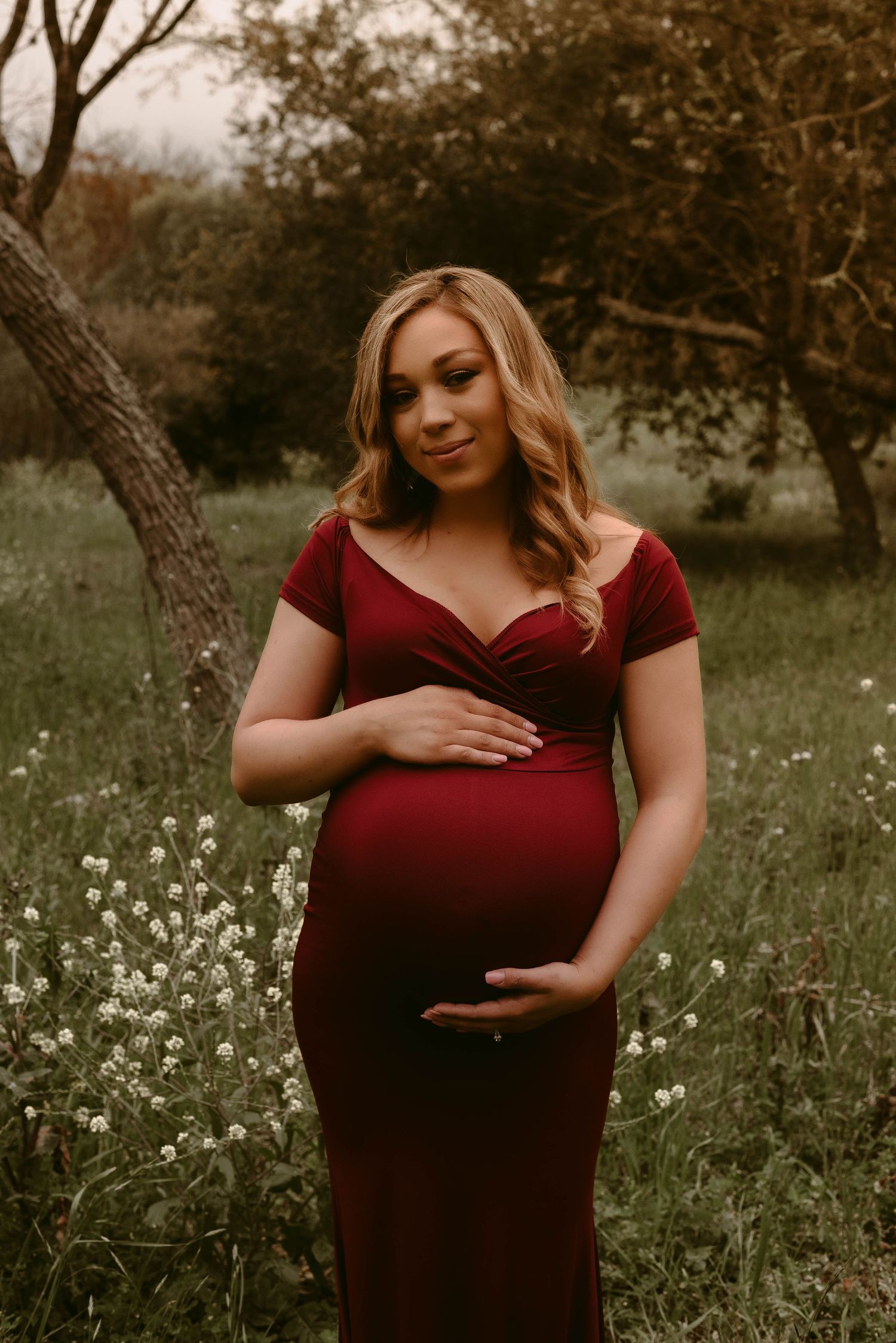 Expectant mother in red dress cradles her belly while standing in a wildflower meadow at sunset.