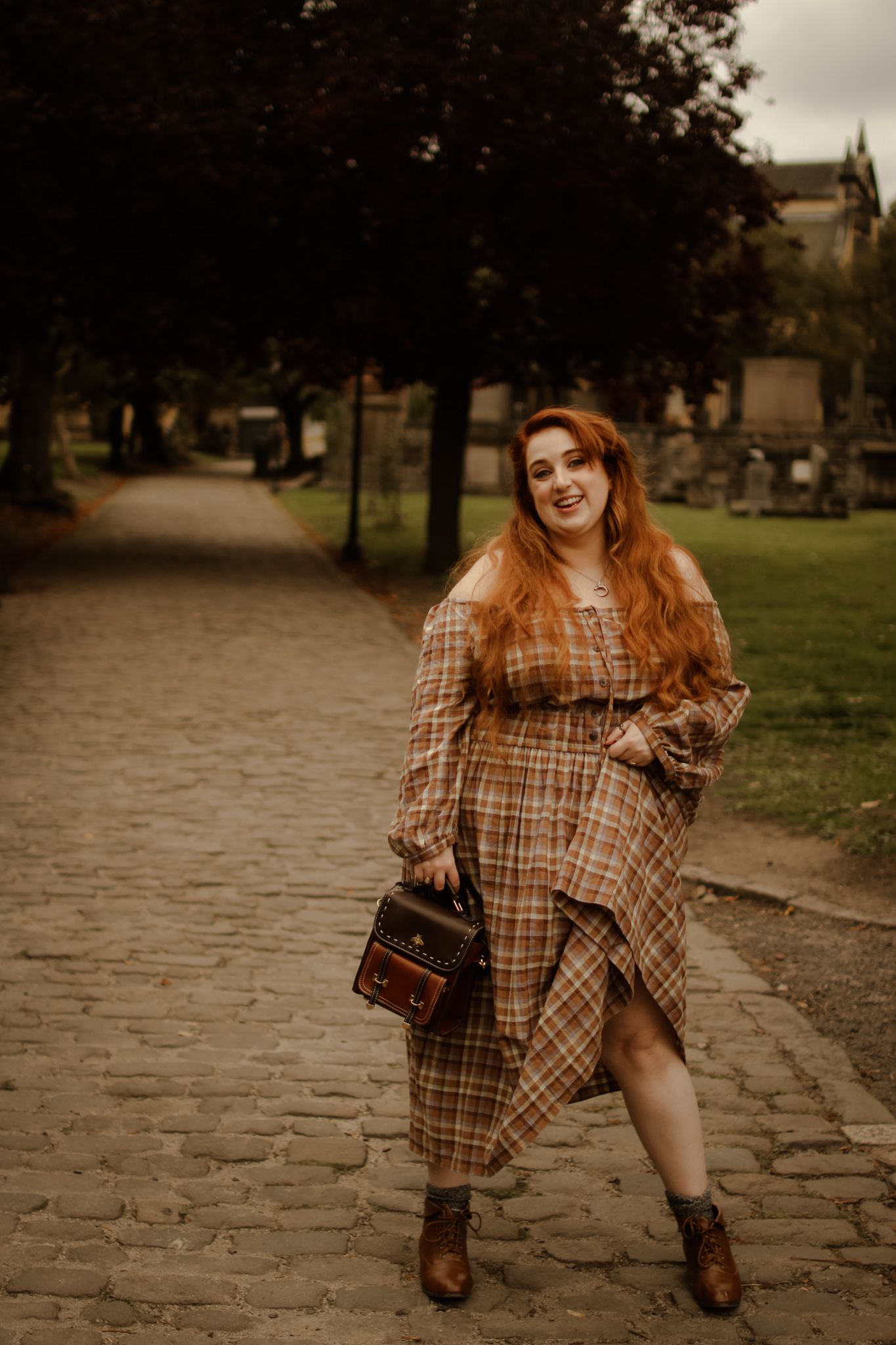 A figure in a plaid dress and boots poses playfully on a cobblestone path during autumn.
