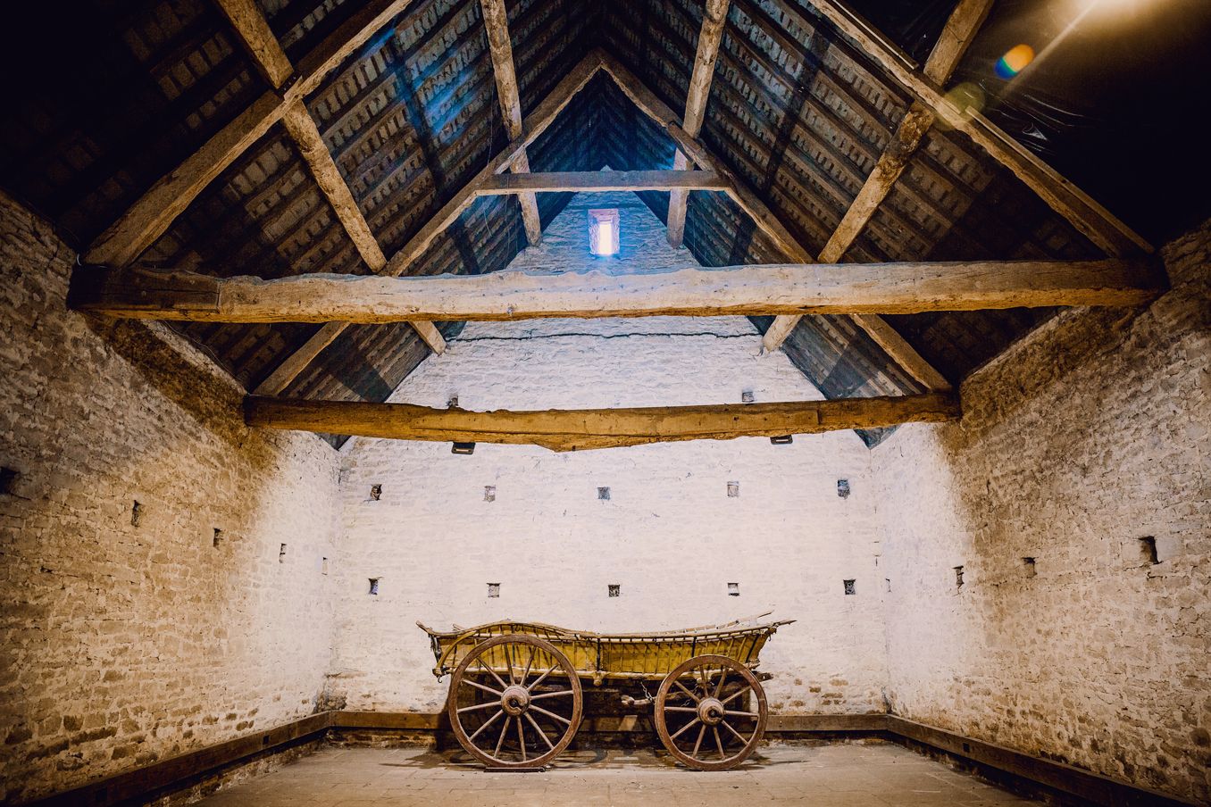 Ancient barn interior with exposed wooden beams and vintage wagon wheels on stone floor.