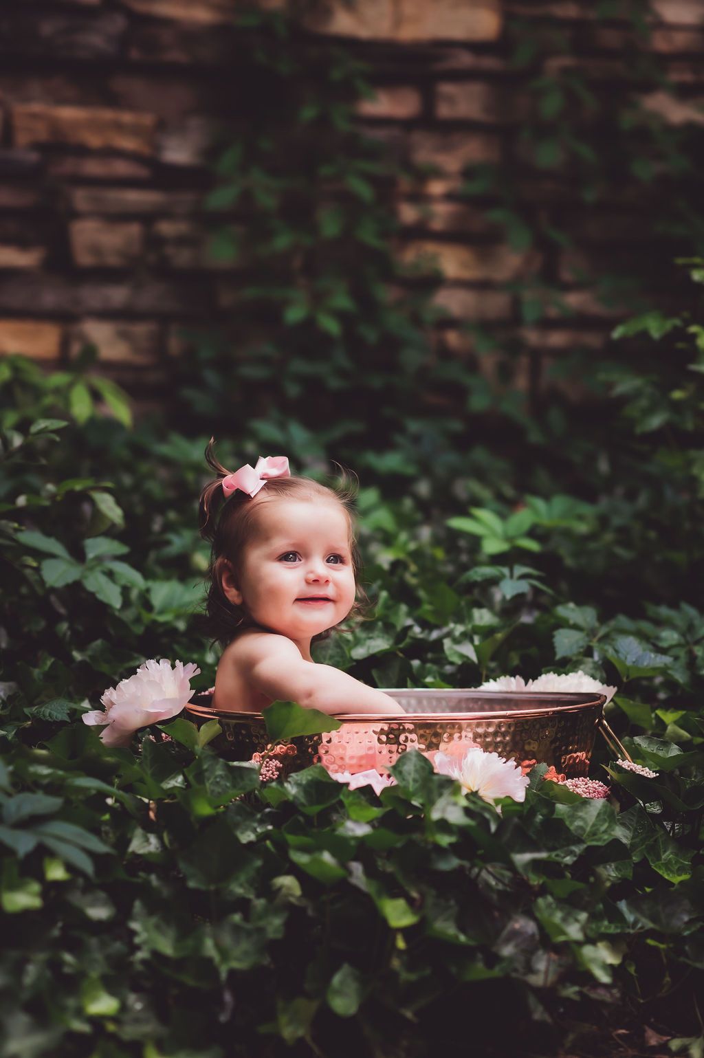 A 7-month-old girl sits in a copper tub set in ivy outside with peonies around the tub for a milk bath photo session