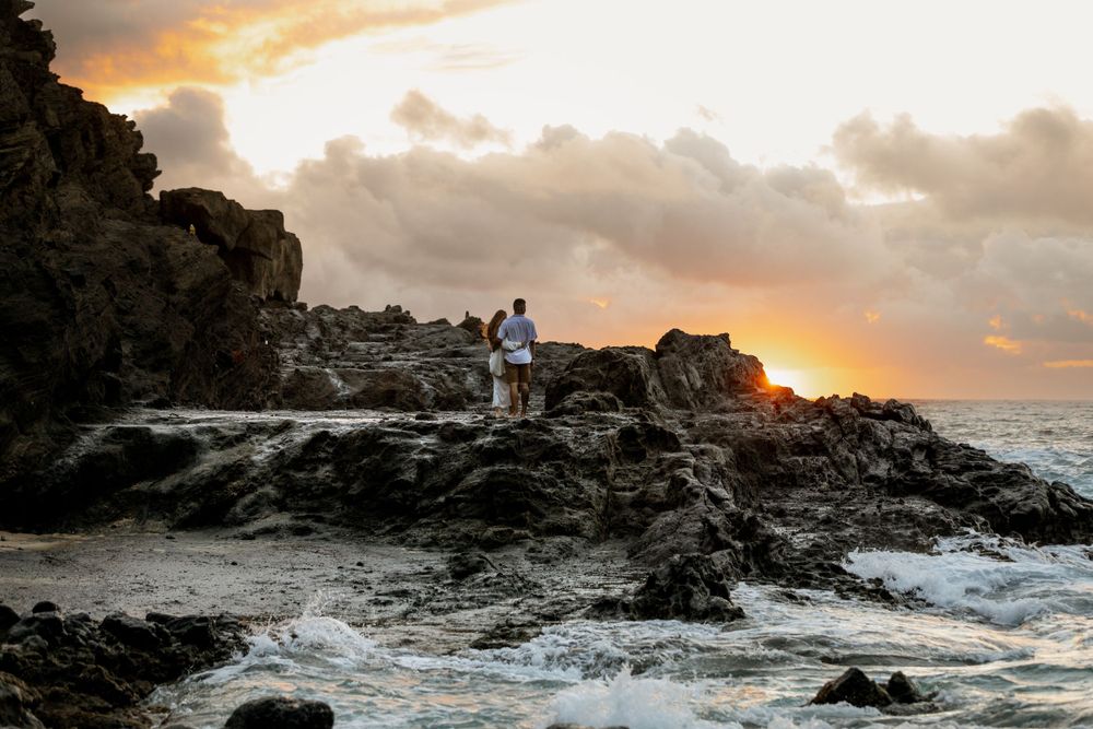 Two figures standing on rocky cliff overlooking ocean at sunset.