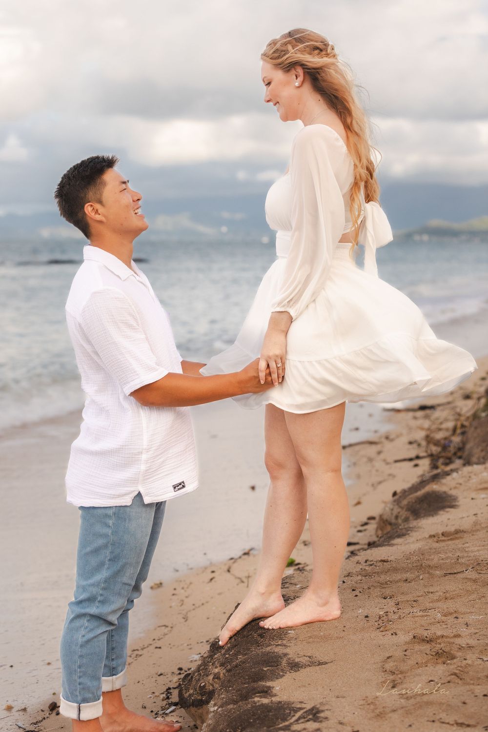 Couple shares playful moment on beach with white dress flowing in ocean breeze.