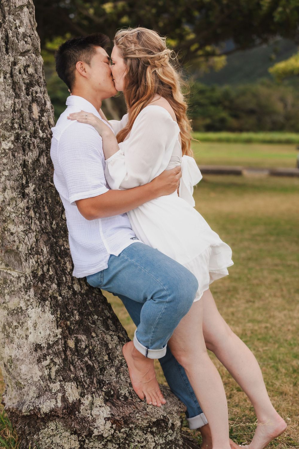 Couple shares romantic kiss against large tree trunk in park setting.