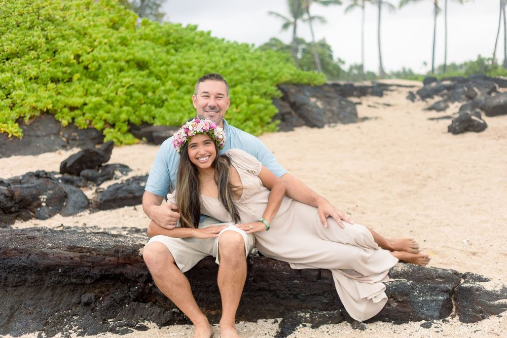 A couple shares a playful moment while sitting on black lava rocks at a tropical beach.