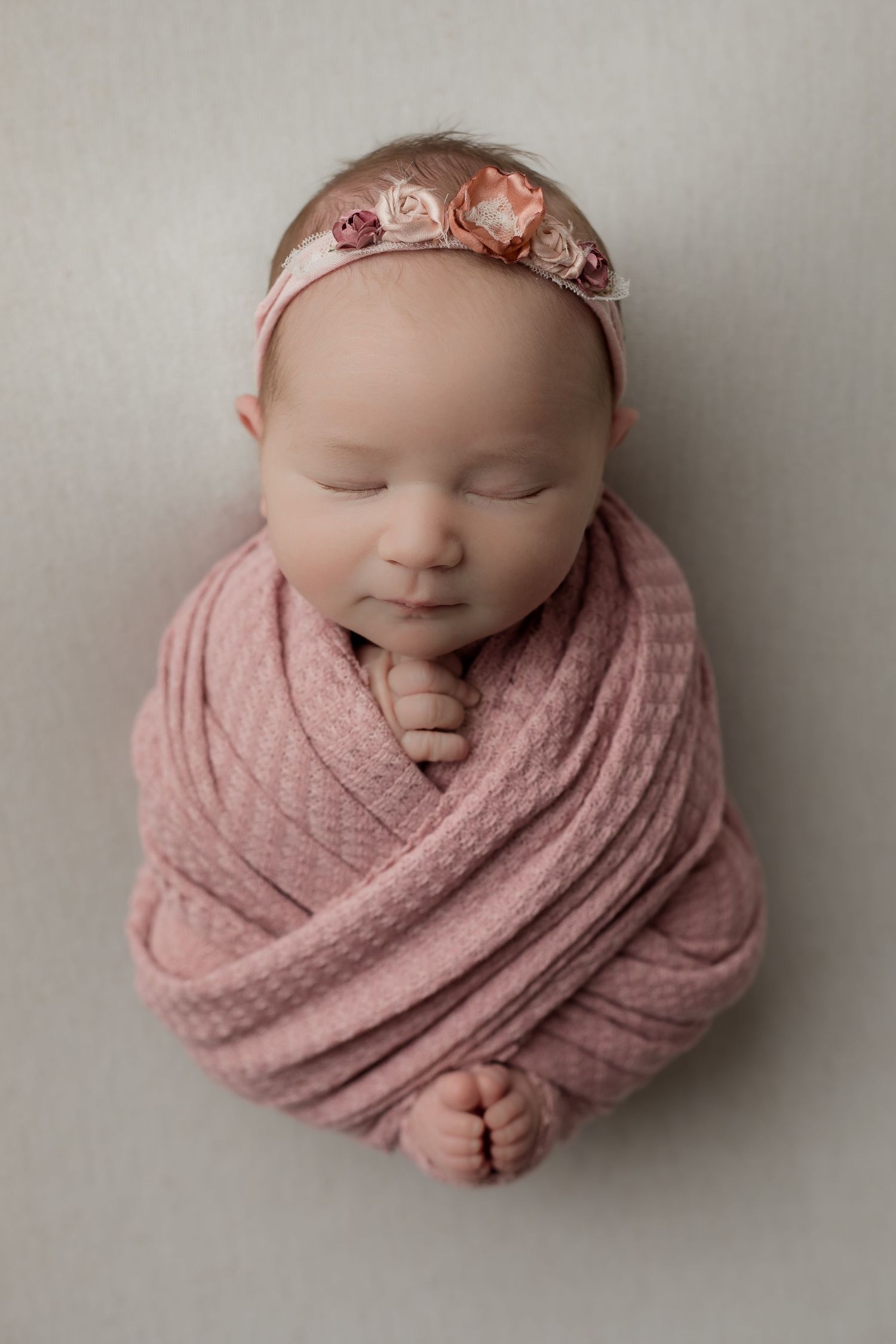 Newborn baby girl posed in a pink swaddle and sleeping on a cream blanket her her studio newborn session.