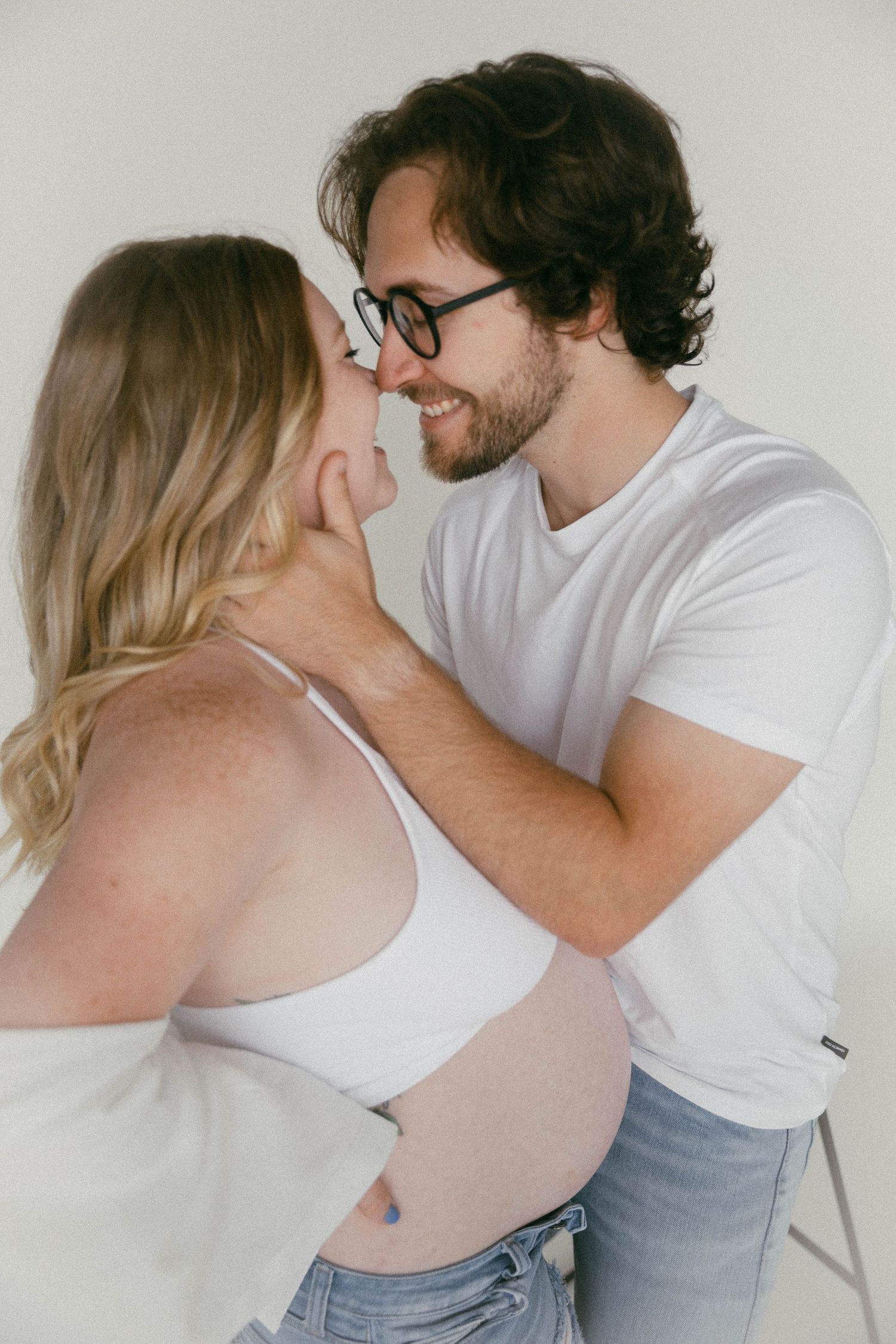 Intimate couple photoshoot against white wall showing tender romantic moments in casual attire.