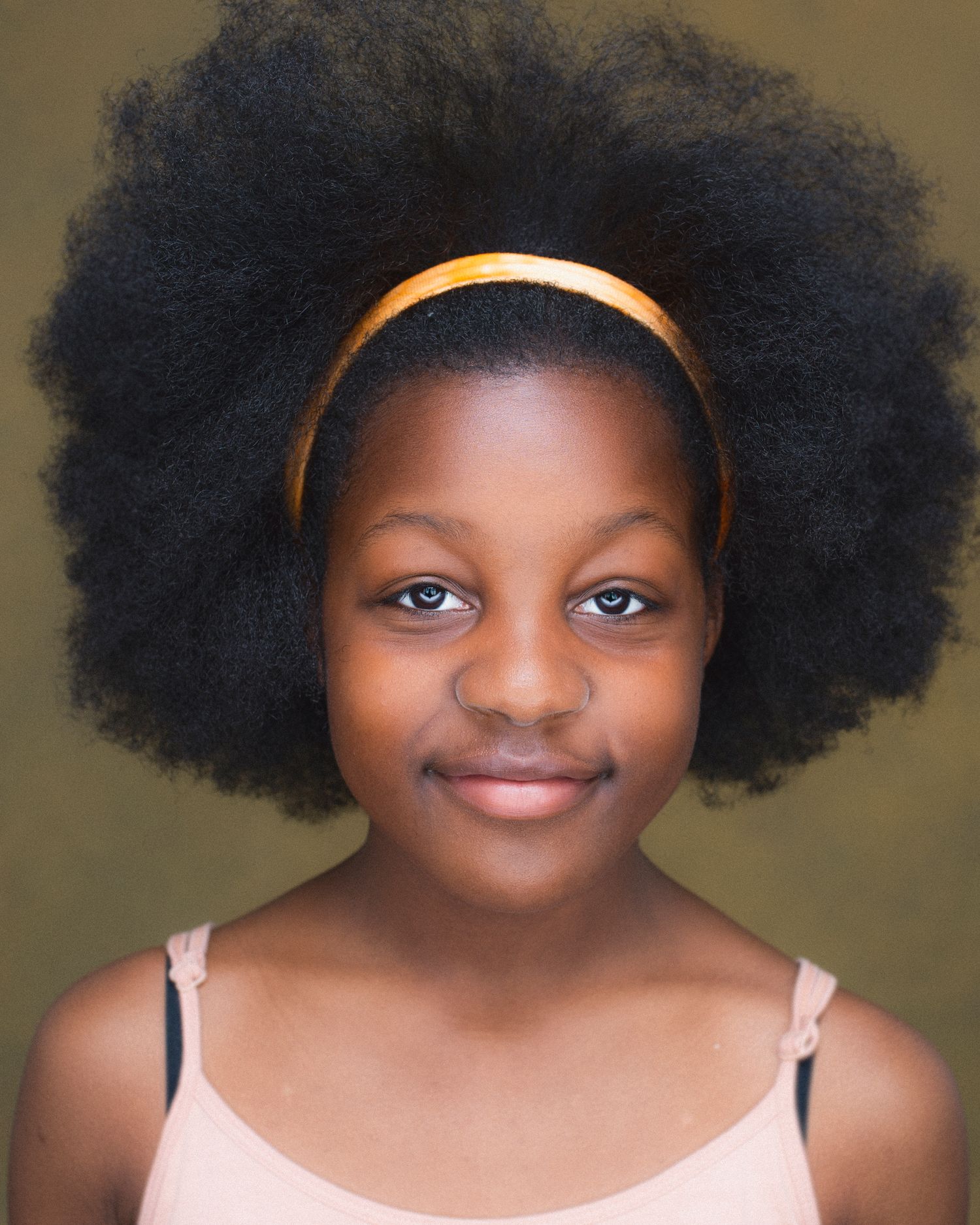 Young girl with natural afro hairstyle wearing gold headband and pink top smiling at camera.