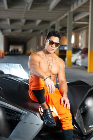 Fitness model wearing orange pants poses on black sports car in urban setting.