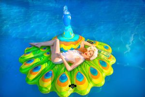 Person floating on a vibrant peacock-inspired pool float in bright blue water.