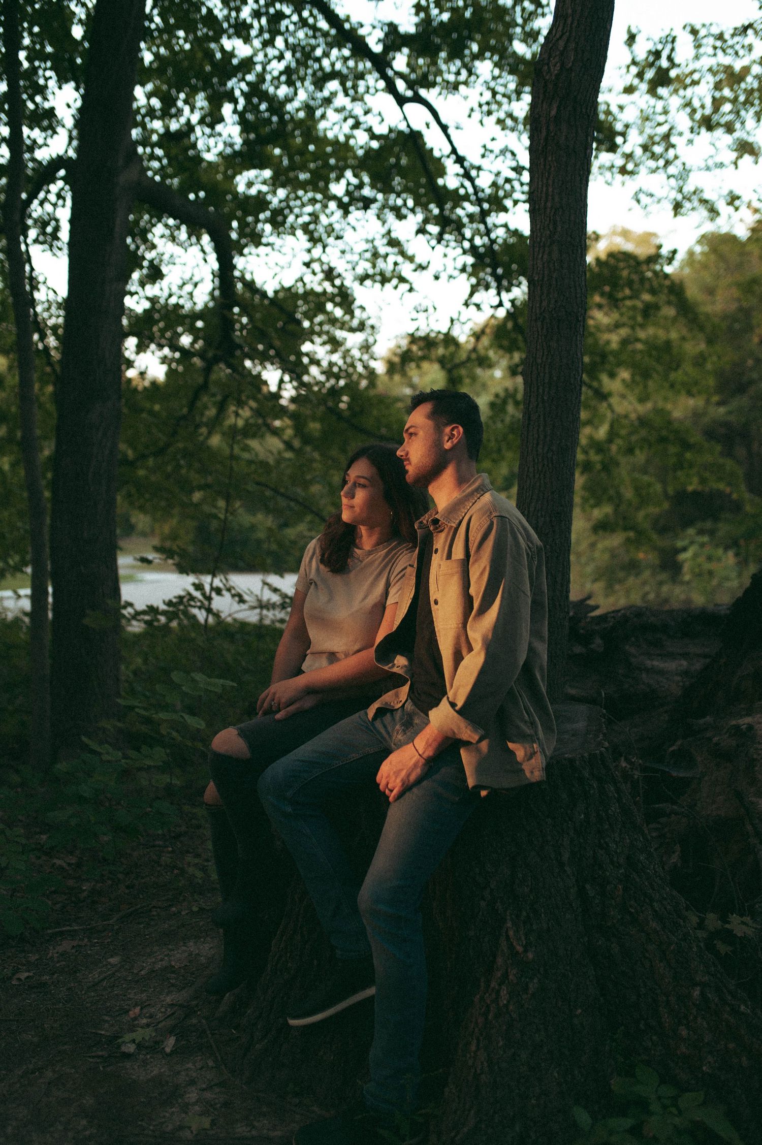 A couple sits together on a tree trunk at sunset in a wooded area.