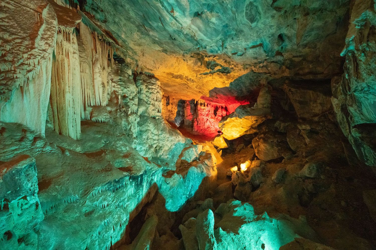 Illuminated cave interior showing colorful rock formations with turquoise and amber lighting.