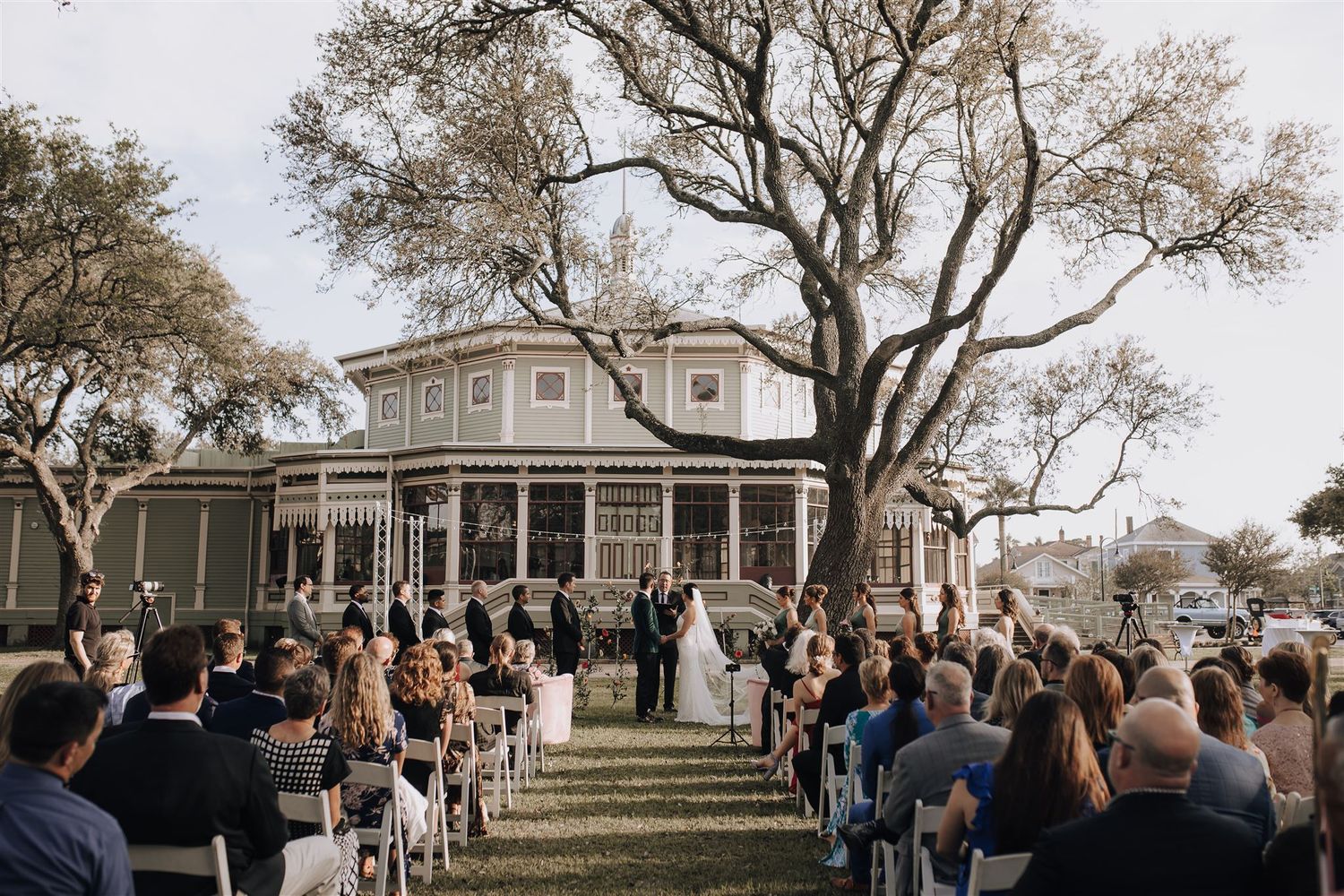 Beautiful outdoor wedding ceremony at a Victorian-style house with guests seated on white chairs.