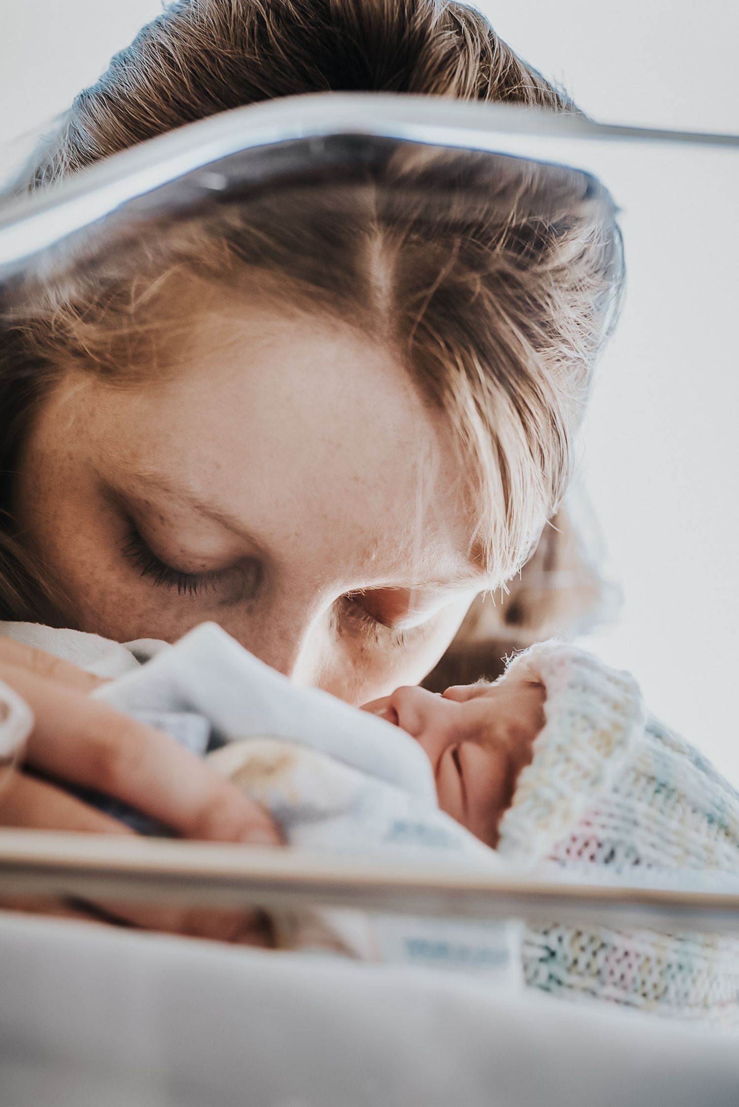 Close up on a mother as she kisses her newborn in a clear basinet.