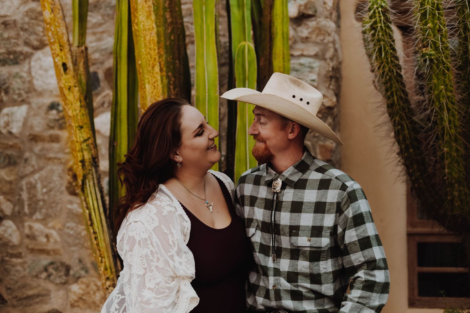 Laughing couple shares a candid moment among desert cacti and rustic stone walls.