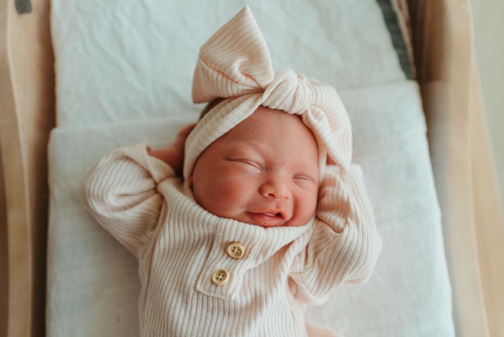 Newborn baby wearing a light pink ribbed outfit with matching bow headband smiles while sleeping.