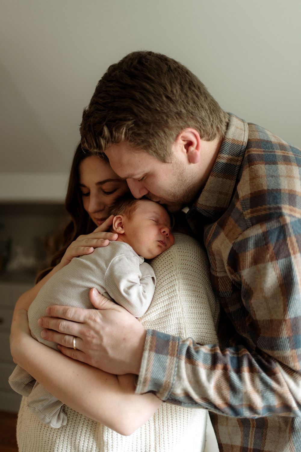 Tender family moment in soft light as parents snuggle with their sleeping newborn baby.