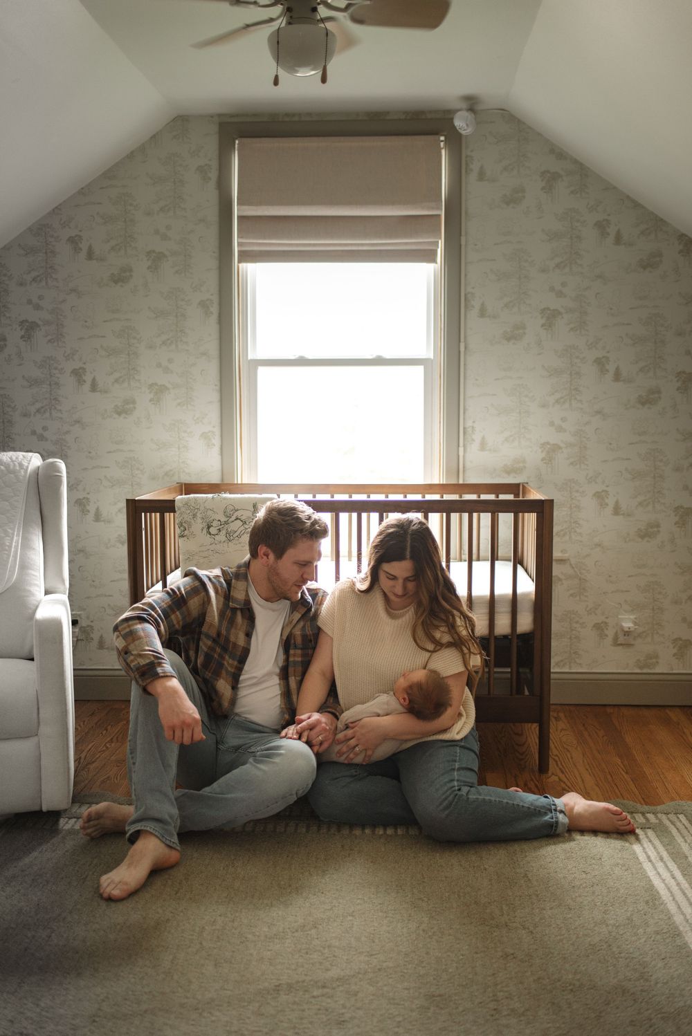 New parents sitting on nursery floor next to wooden crib sharing a special moment.