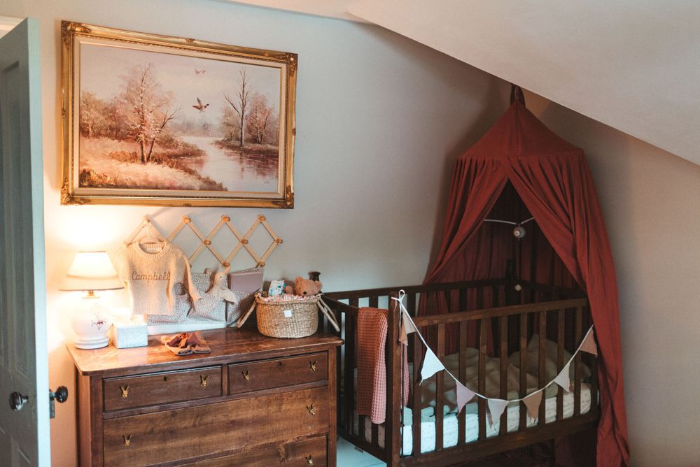 Cozy nursery corner with vintage dresser, landscape art and burgundy canopy over wooden crib.