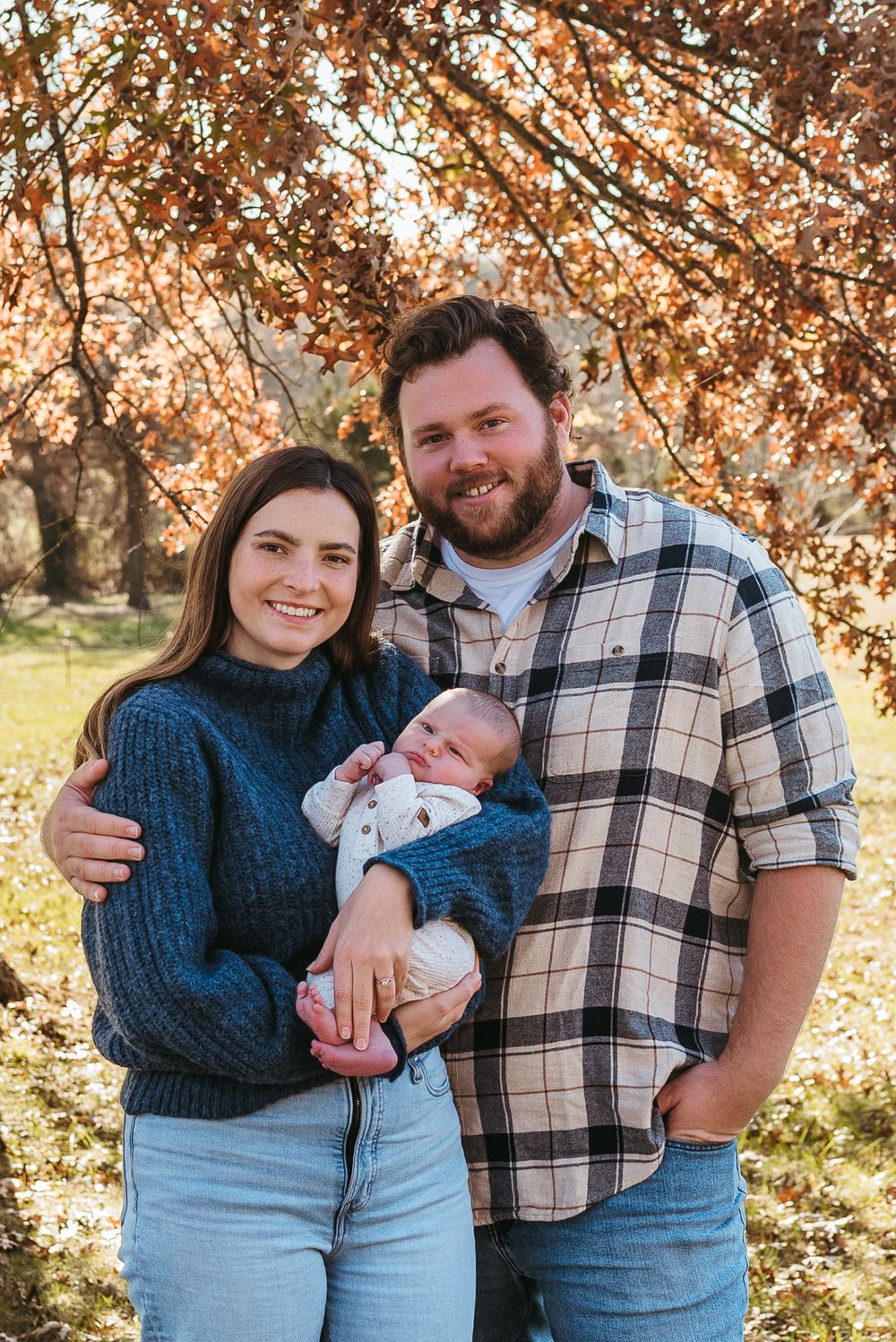 A cozy family portrait outdoors with autumn foliage featuring a couple holding their newborn baby.