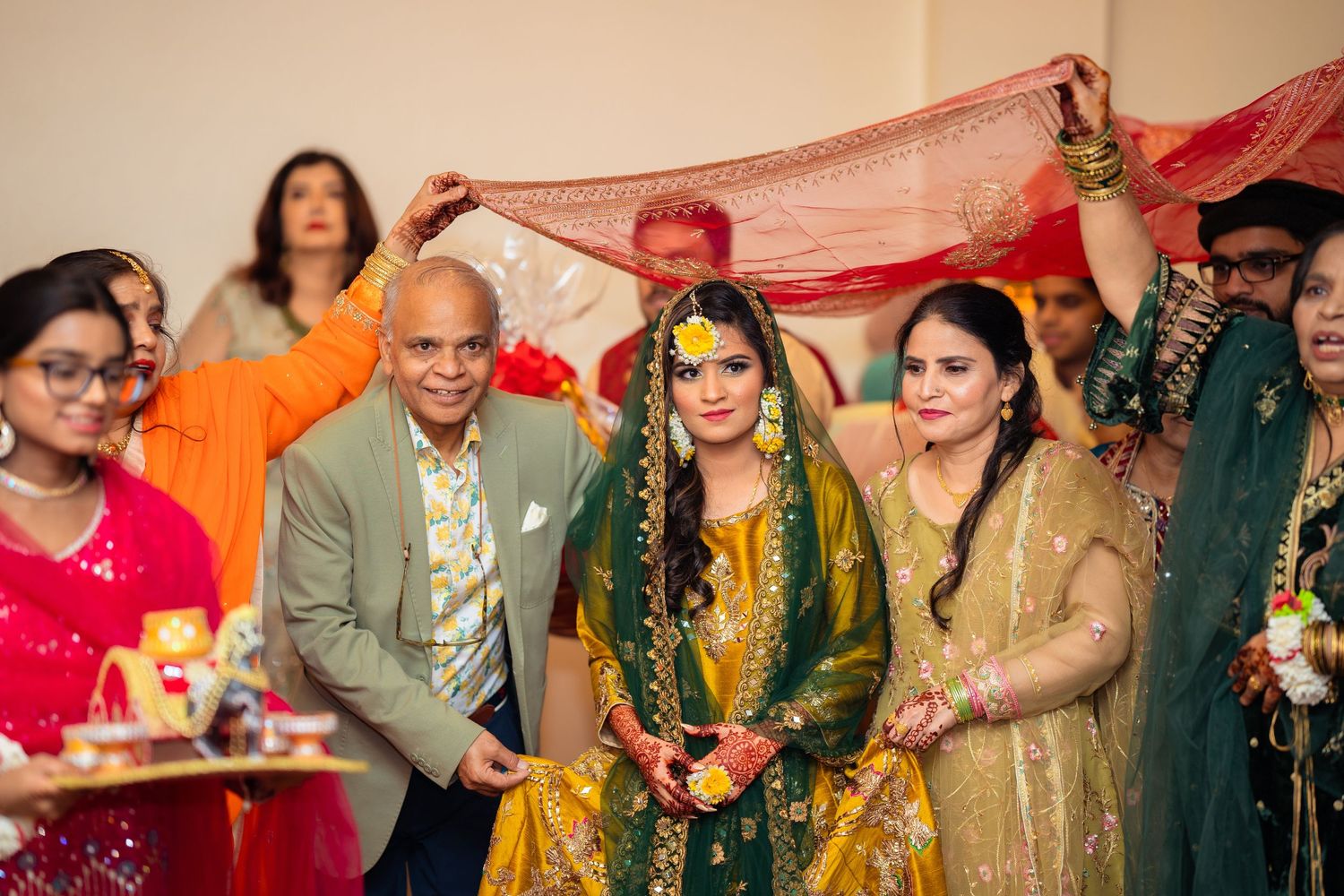 Wedding ceremony guests gather around bride under red dupatta during traditional South Asian celebration.