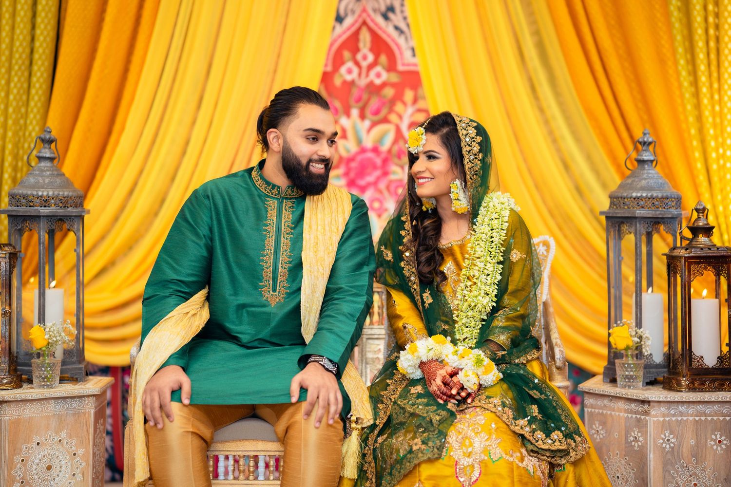 Couple in matching green traditional outfits pose against yellow backdrop with decorative lanterns.