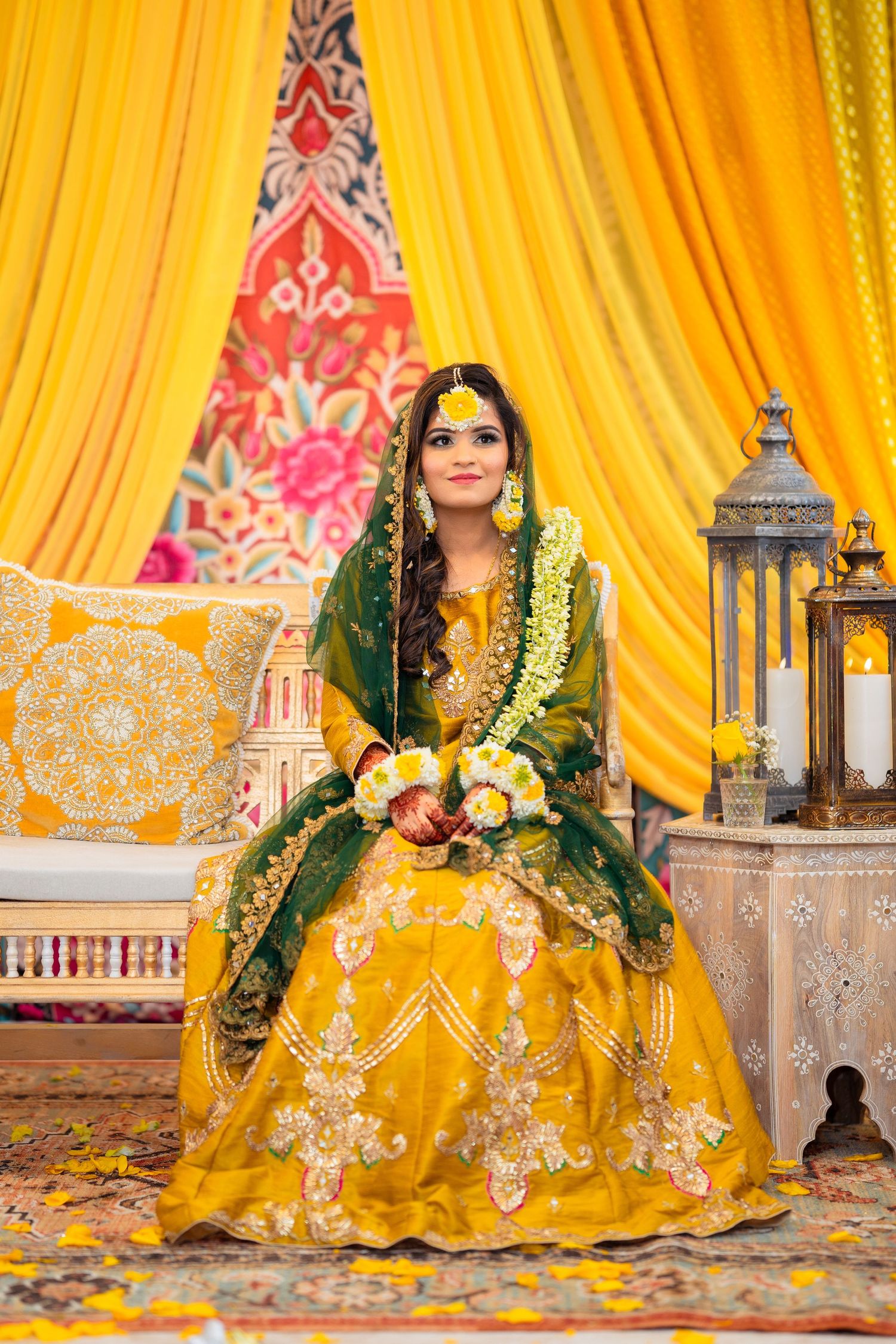 Bride in yellow and green lehenga poses on ornate chair against yellow drapery with decorative lanterns.