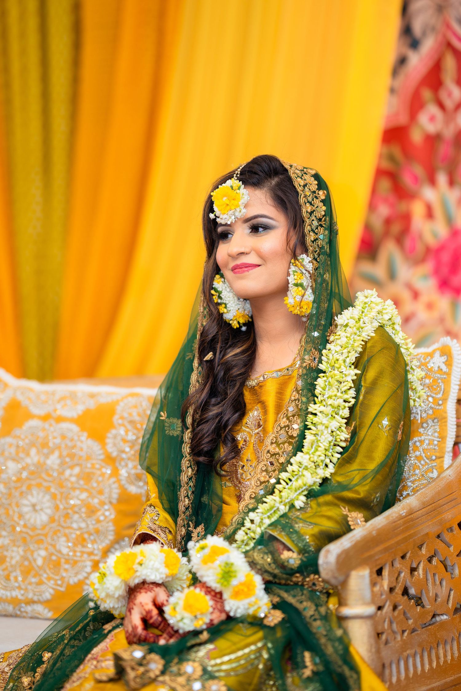 Bride in yellow and green traditional dress adorned with floral jewelry sits on decorated chair.