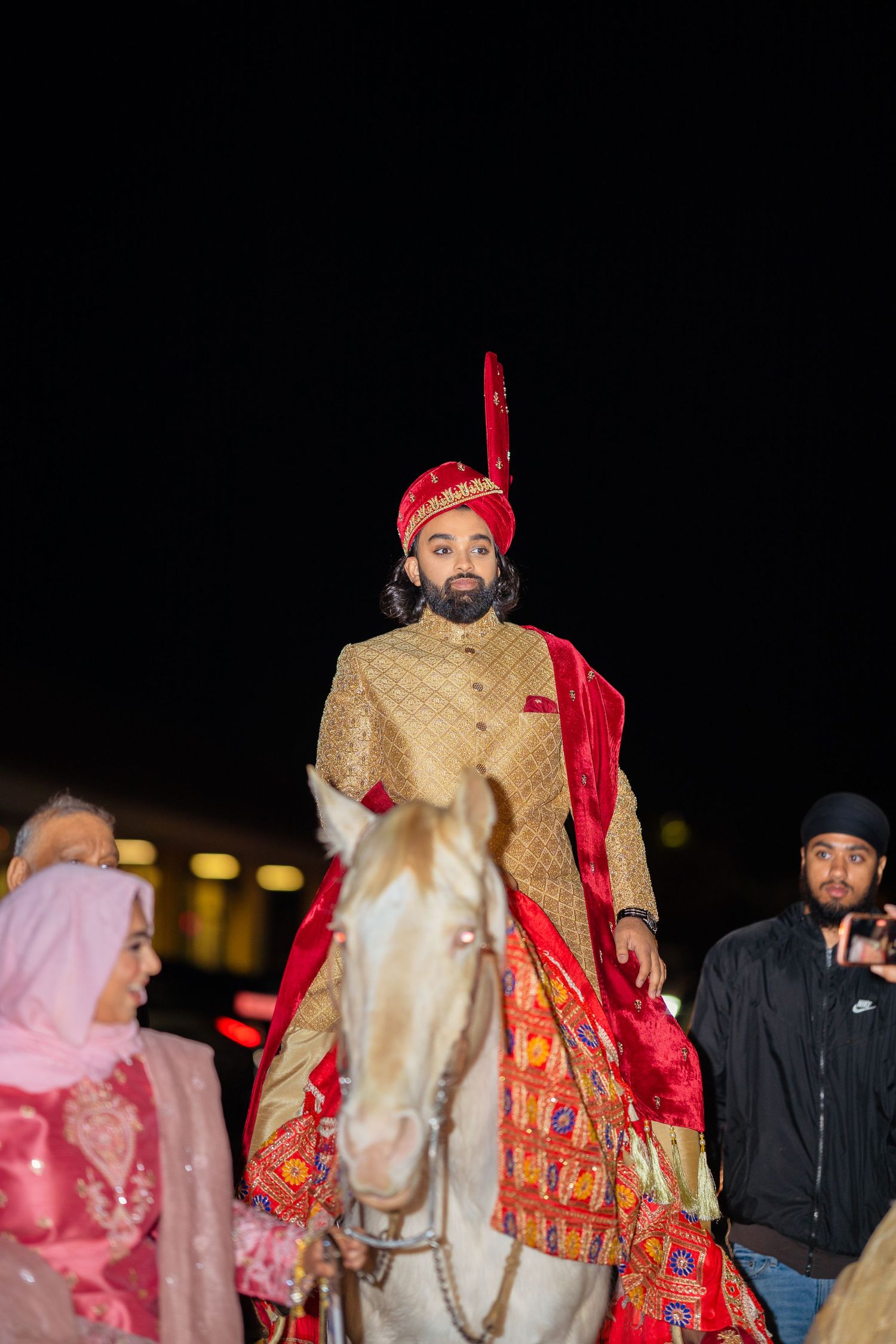 Groom in traditional South Asian attire rides white horse during night baraat procession.