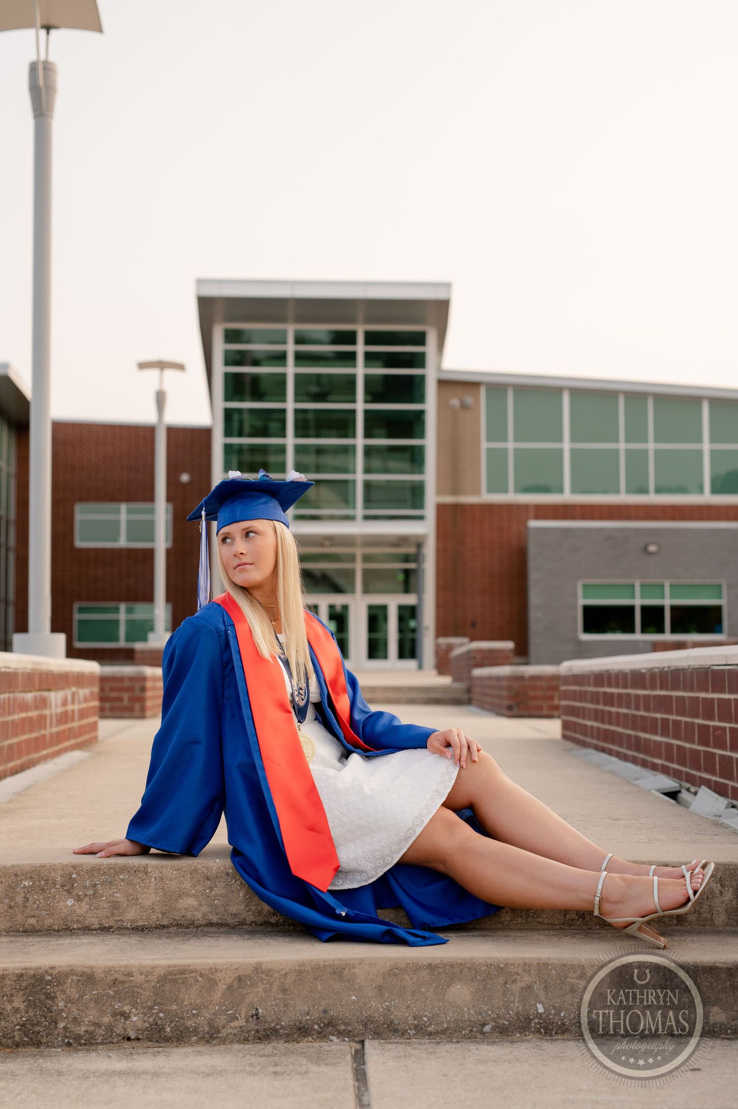Graduate in blue regalia and red tie poses on brick steps outside modern educational building.