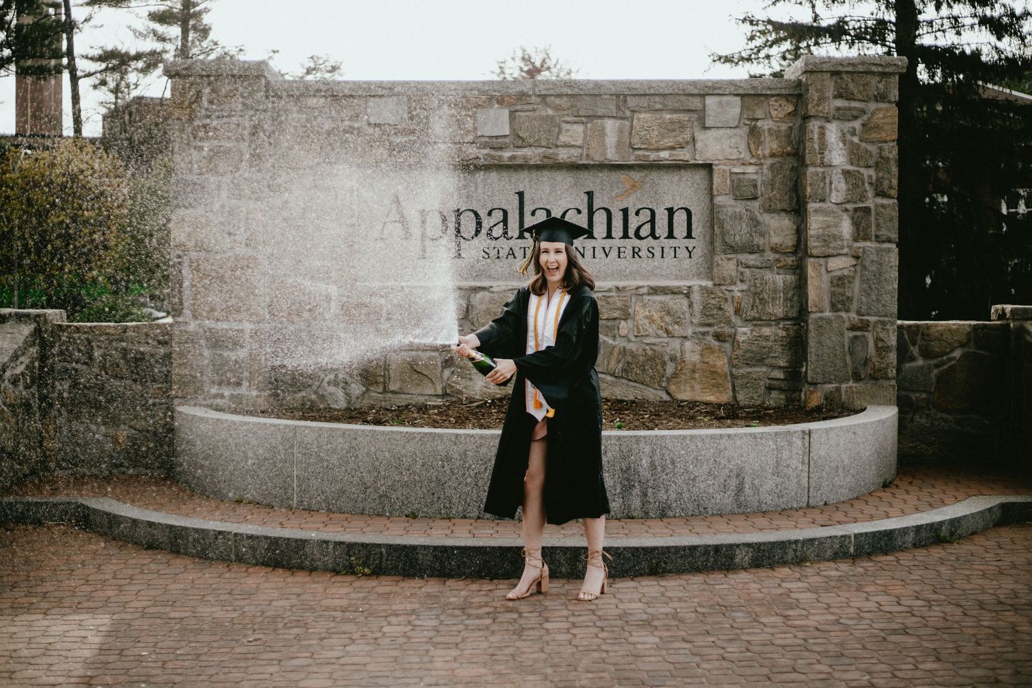 Graduate standing by Appalachian university stone entrance sign wearing formal attire.