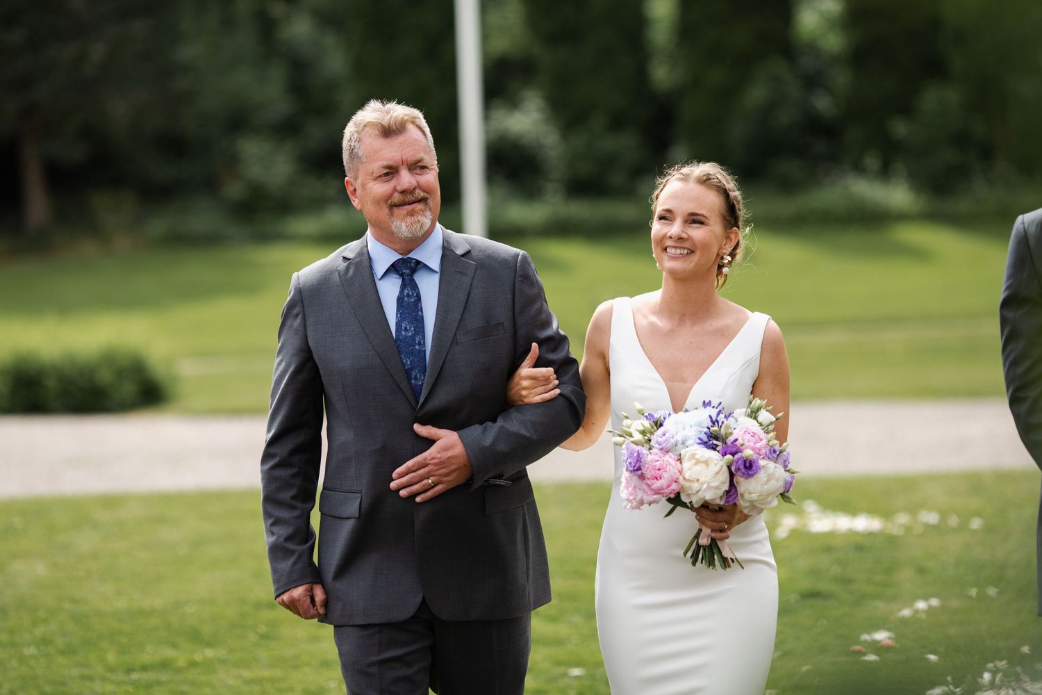 Joyful walk down outdoor wedding aisle with elegant white dress and purple bouquet.