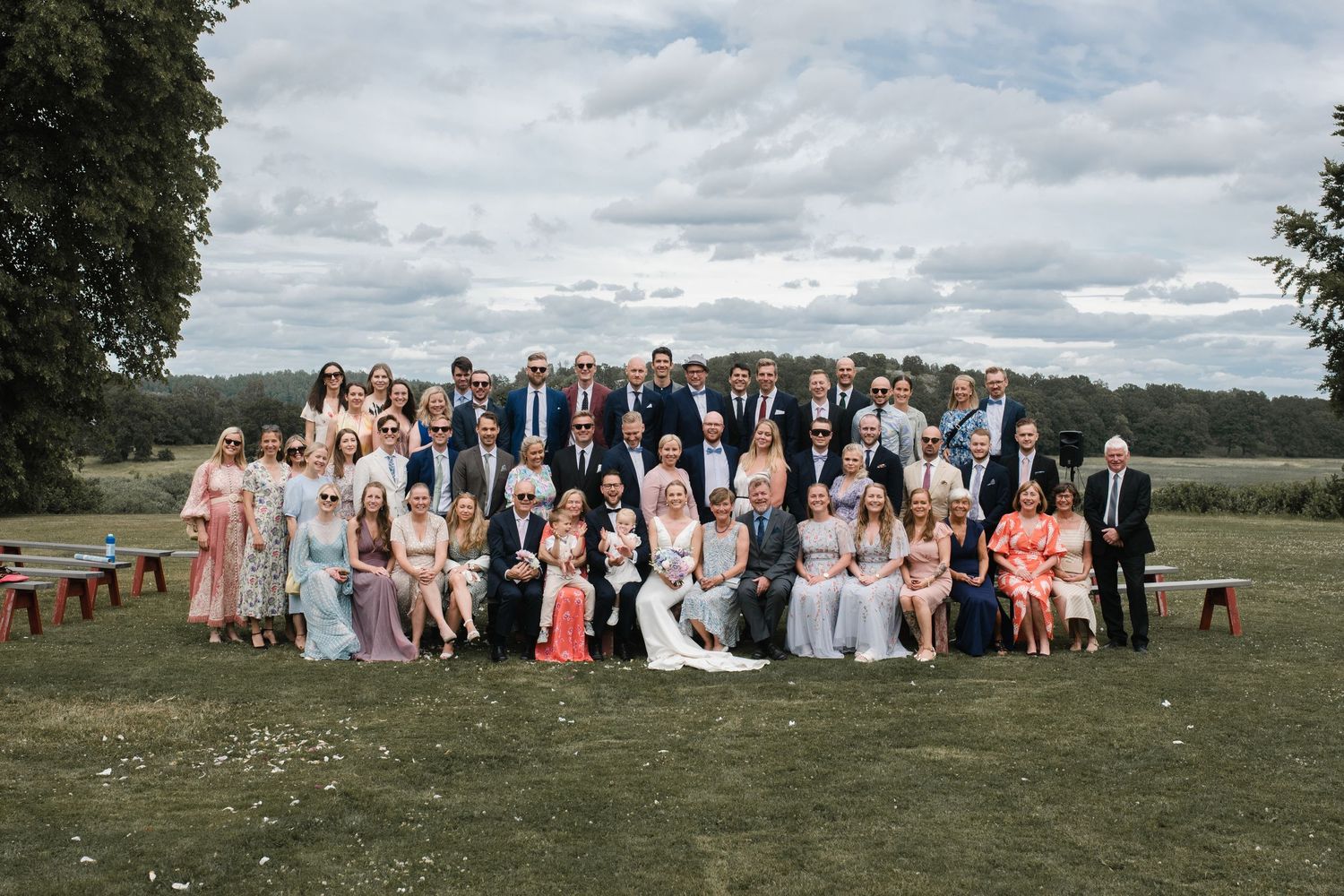 Large wedding group photo of guests and wedding party gathered outdoors on grassy hill.
