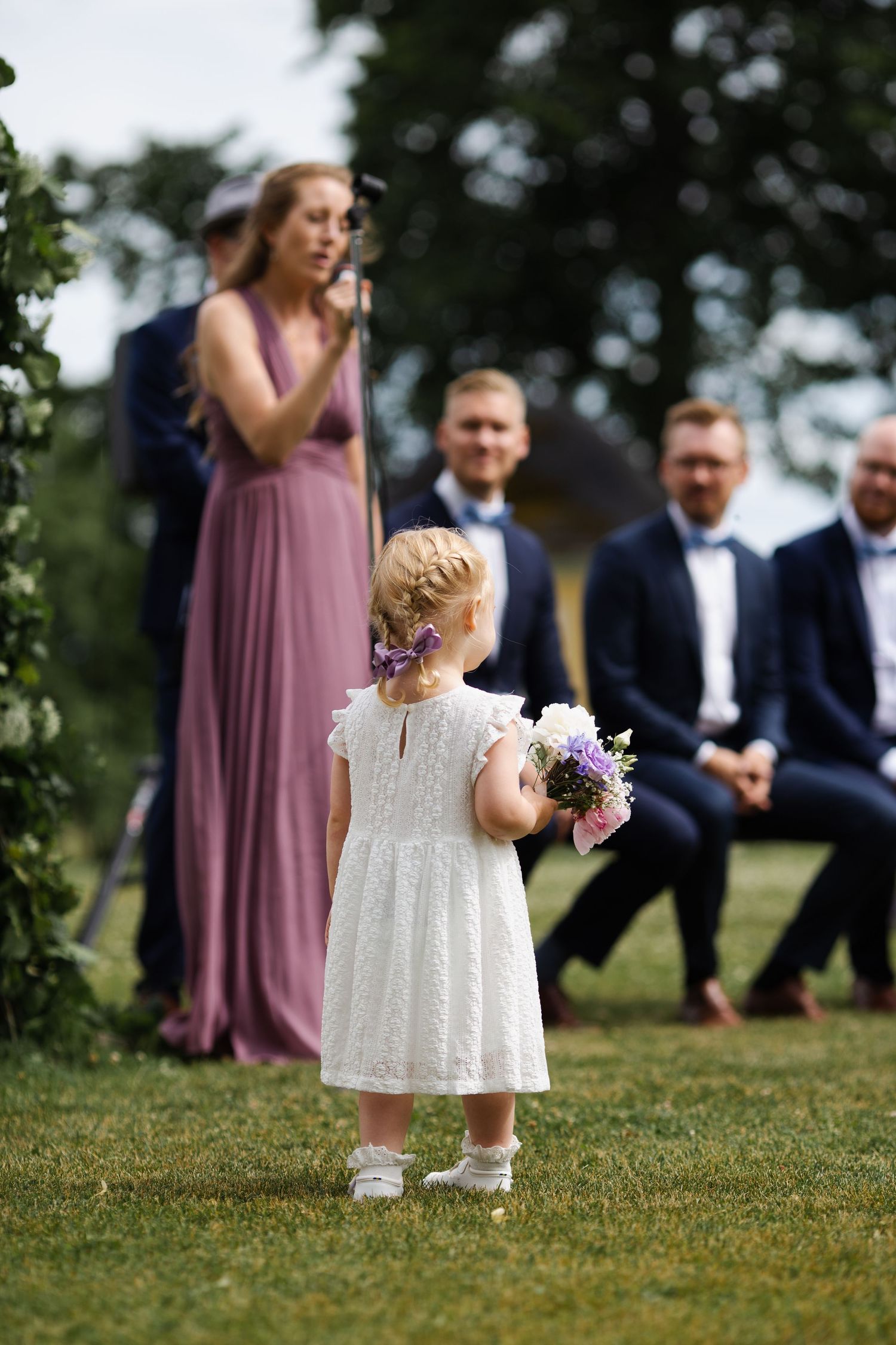 Small flower girl in white dress holds bouquet during outdoor wedding ceremony.