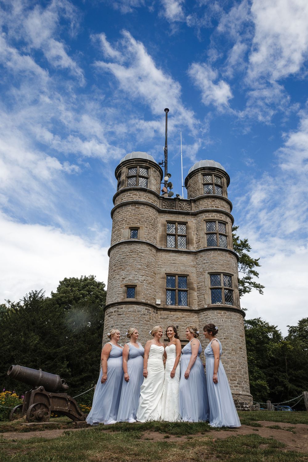 Wedding party poses in front of a stone castle tower with turrets under bright blue skies.