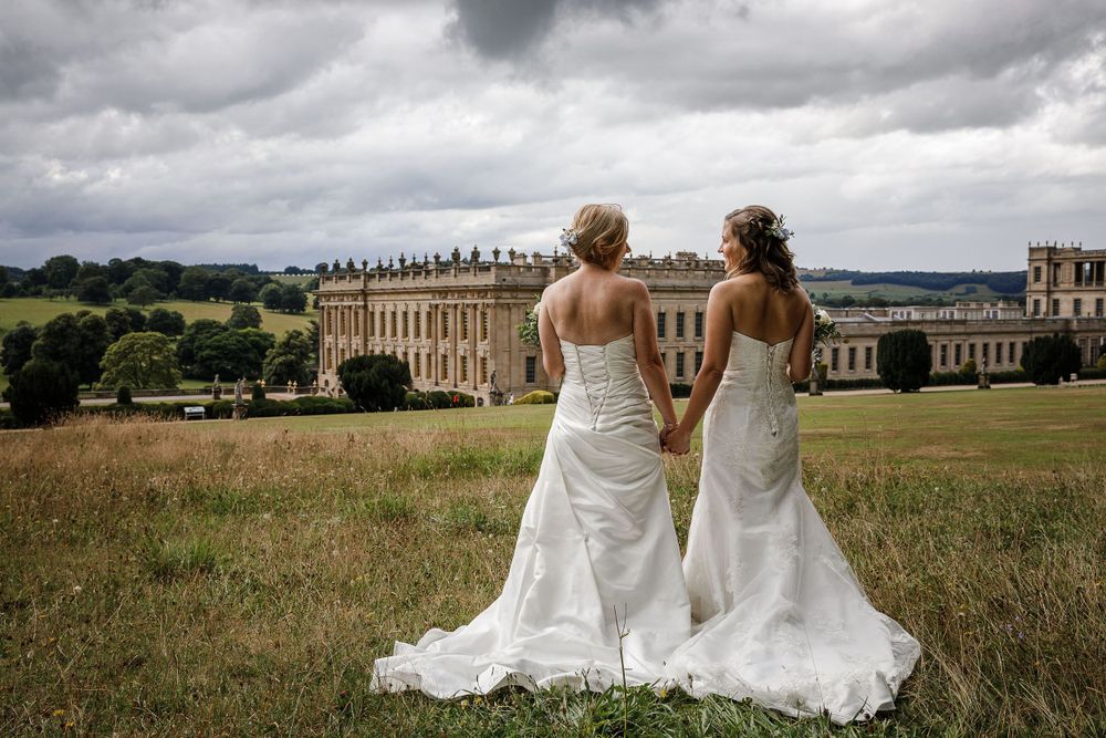 A couple in white wedding dresses poses in a scenic field with a grand manor house in the background.